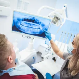 Lekarz w masce i rękawicach pokazuje pacjentowi zdjęcie rentgenowskie szczęki na monitorze podczas wizyty w gabinecie.