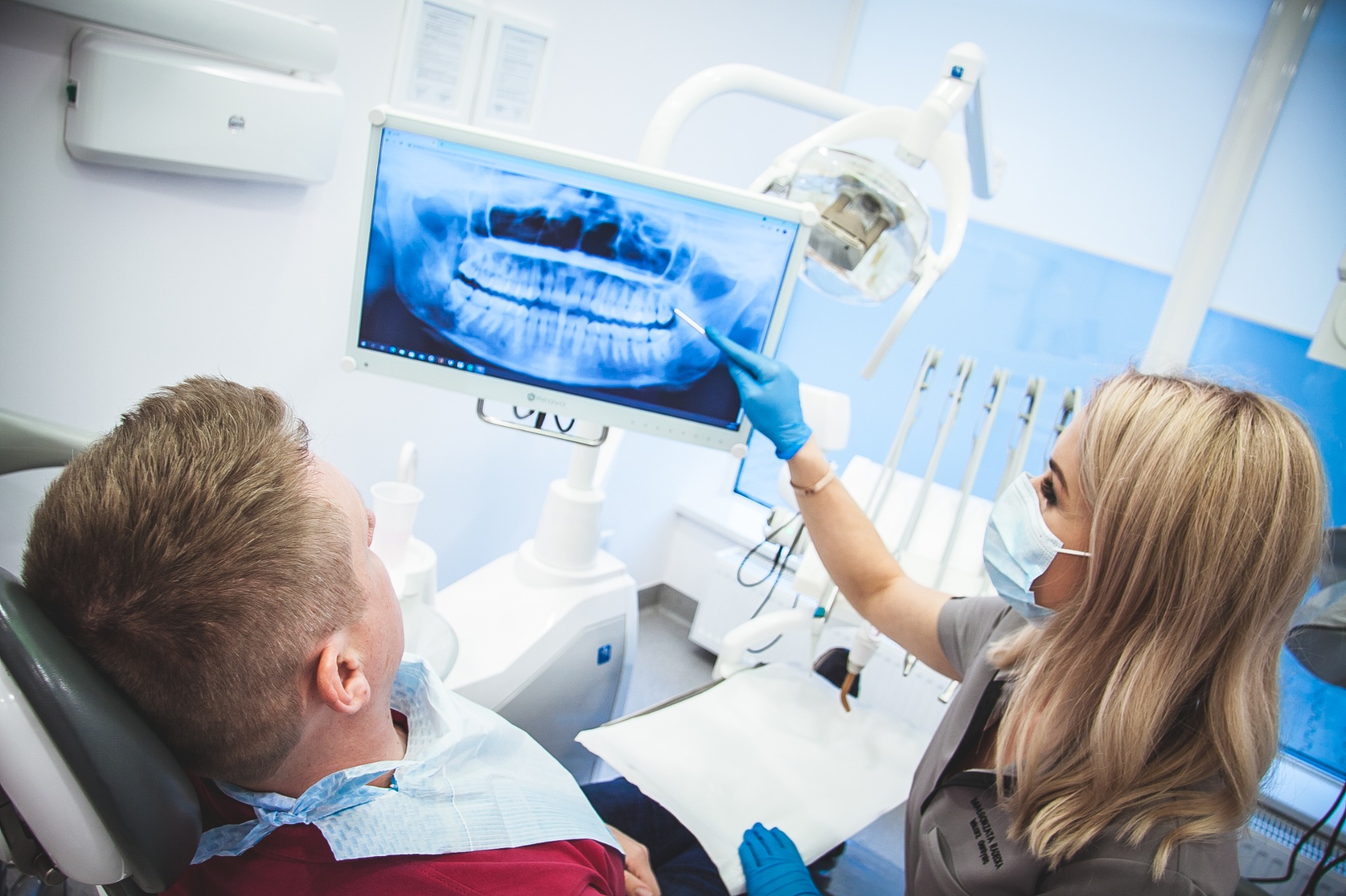 Lekarz w masce i rękawicach pokazuje pacjentowi zdjęcie rentgenowskie szczęki na monitorze podczas wizyty w gabinecie.