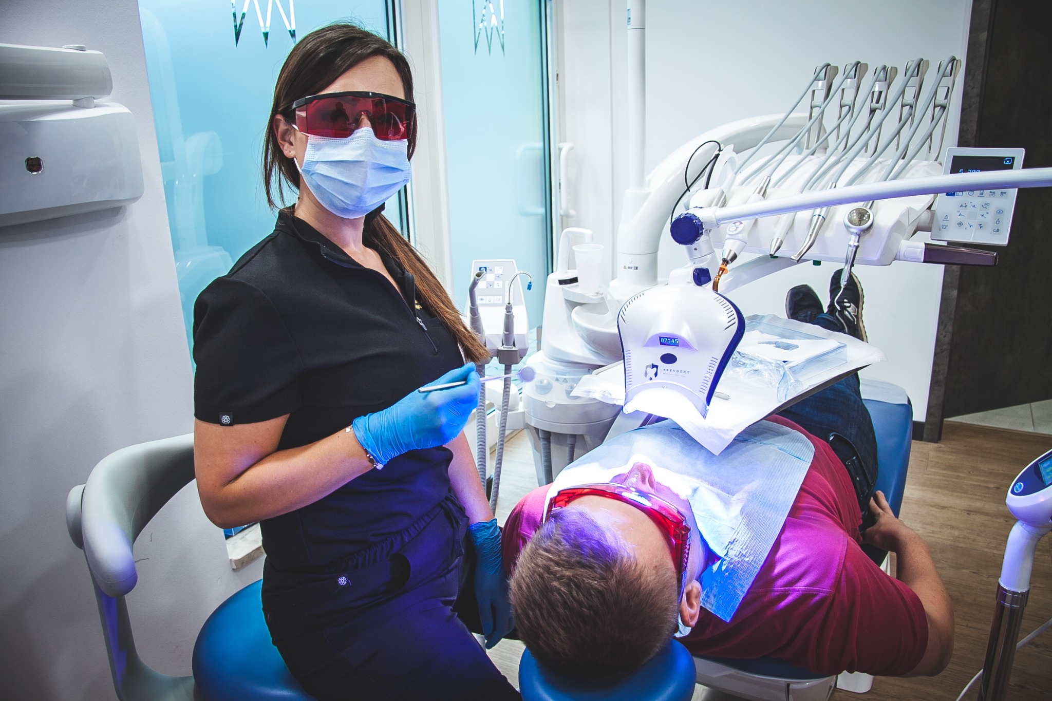 Stomatolog w okularach ochronnych i maseczce, trzymająca narzędzie dentystyczne, stoi obok pacjenta leżącego na fotelu dentystycznym pod lampą polimeryzacyjną.