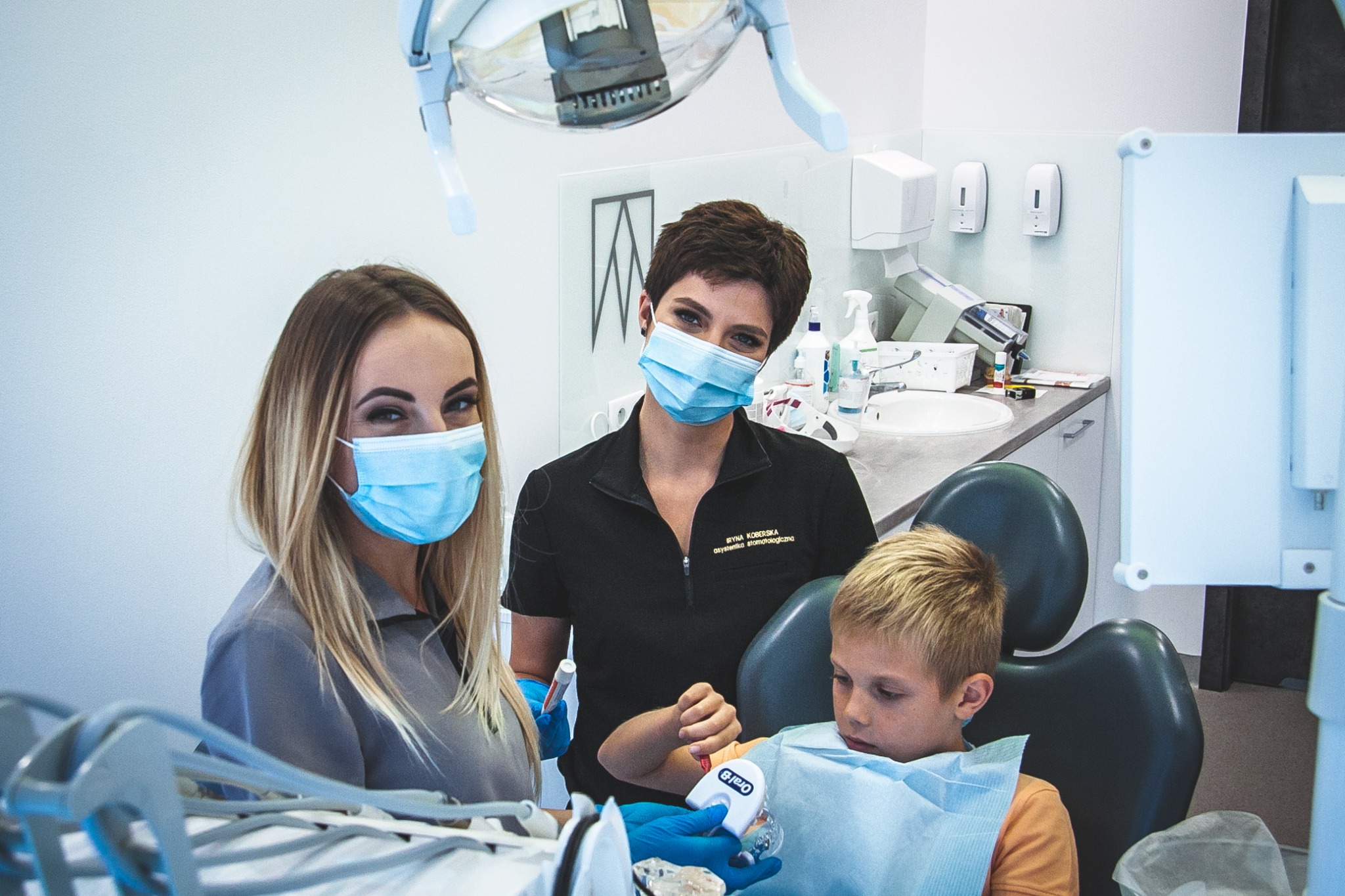 Dwaj lekarze w maskach i rękawiczkach przeprowadzają badanie jamy ustnej u chłopca w gabinecie z nowoczesnym sprzętem, widoczny unit dentystyczny i lampa zabiegowa.