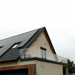 Nowoczesny dom z balkonem zabezpieczonym białą, metalową konstrukcją, panele słoneczne na dachu, widok z dołu na tle pochmurnego nieba.