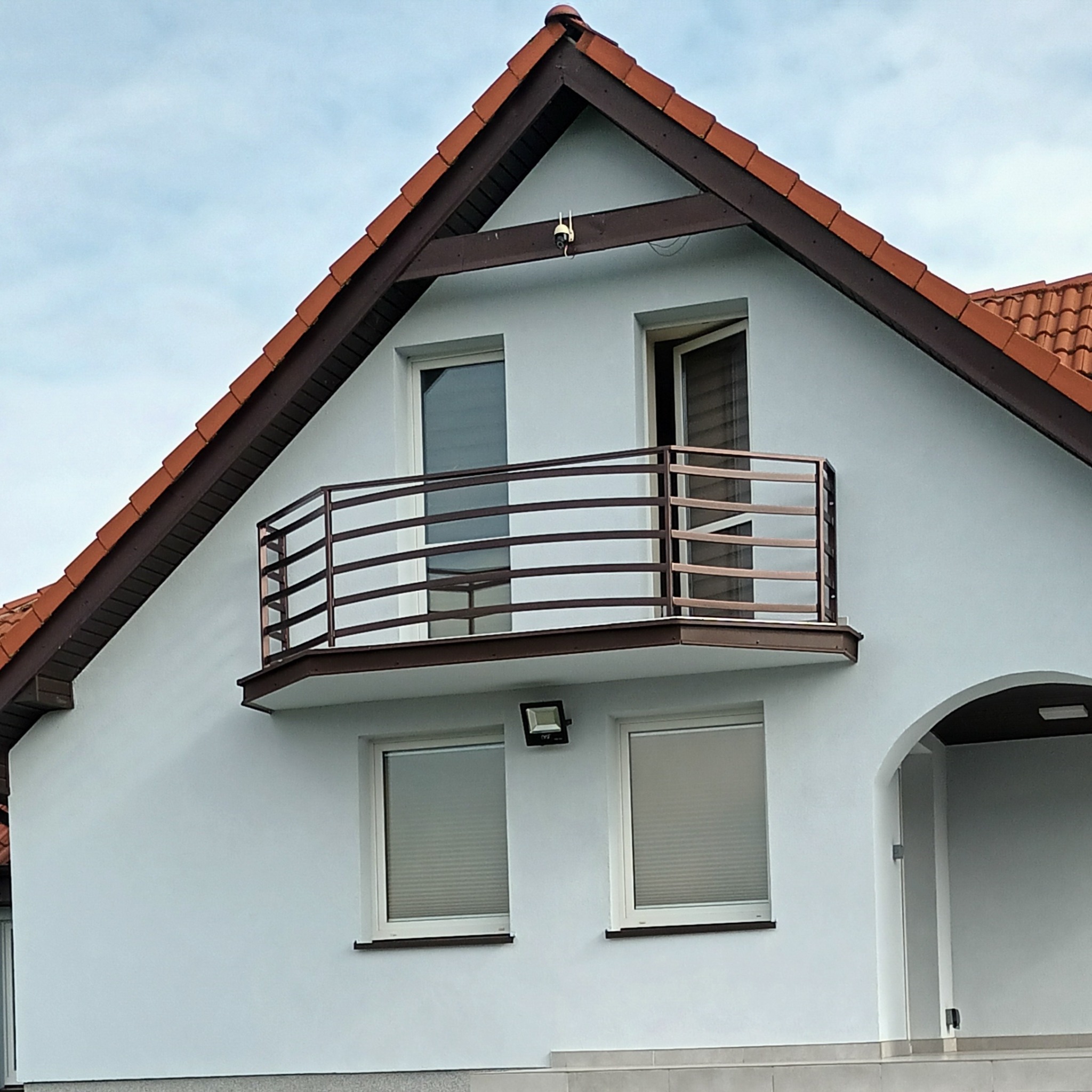 Elewacja domu z balkonem zabezpieczonym poziomą, brązową konstrukcją z metalowych prętów; widoczny fragment dachu z czerwoną dachówką, okna z żaluzjami i lampa na ścianie.