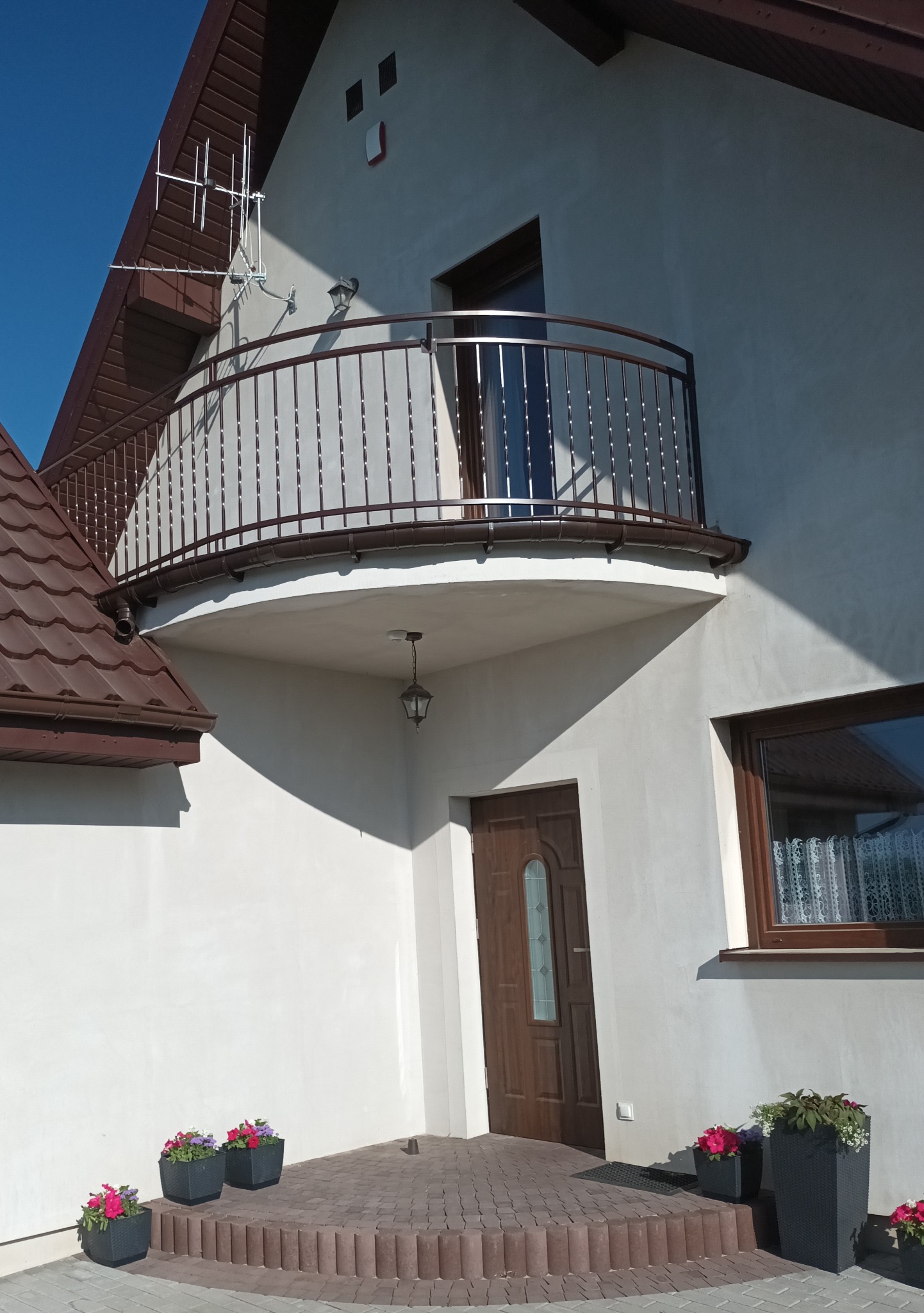 Balkon z brązową, metalową balustradą o zaokrąglonym kształcie, z widocznymi skręconymi elementami pionowymi, umieszczony na białym budynku z brązowym dachem i oknami, z kwiatami w donicach...
