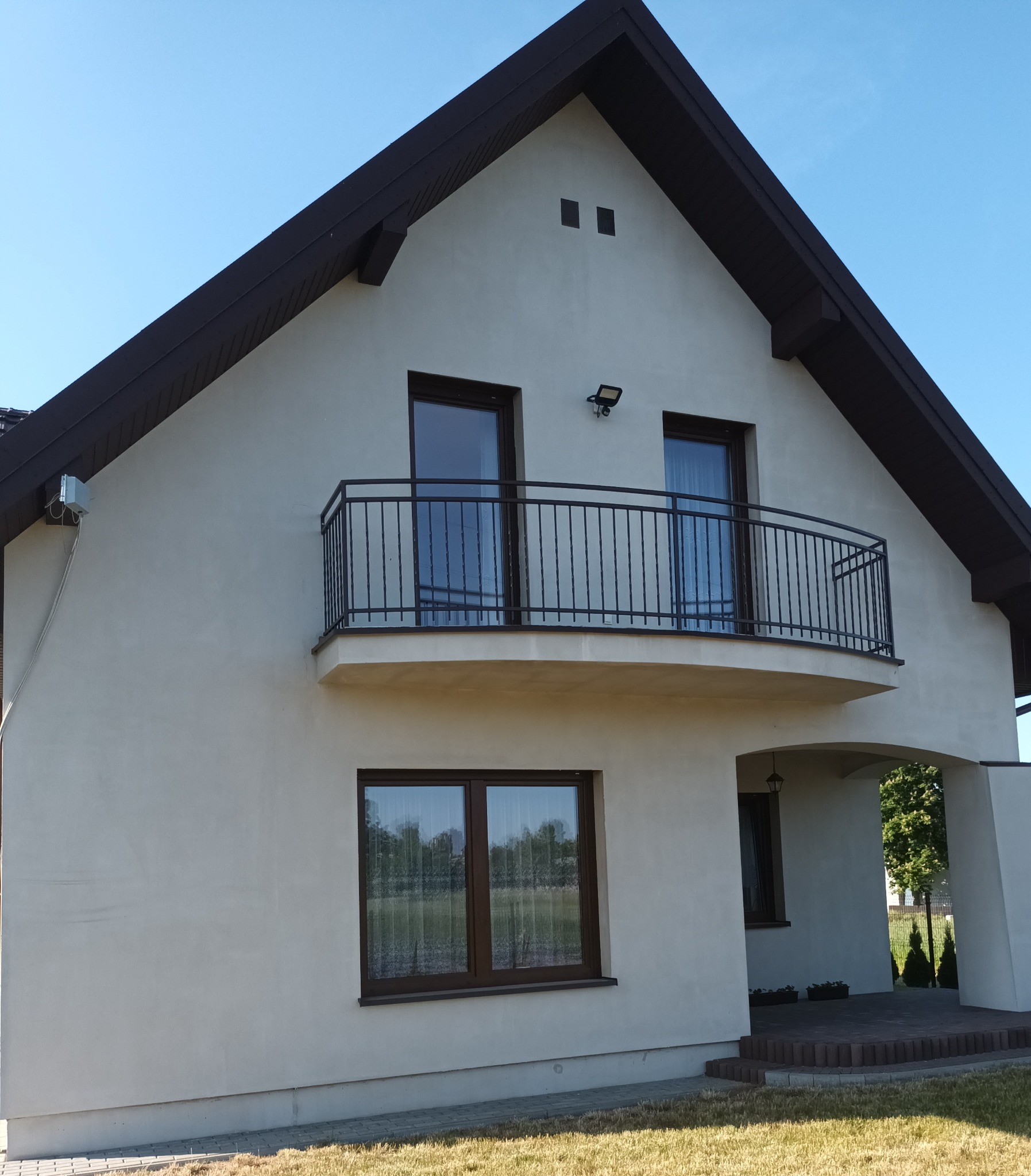 Dwupiętrowy dom z balkonem zabezpieczonym czarną, metalową balustradą, brązowe ramy okienne, jasna elewacja, błękitne niebo.