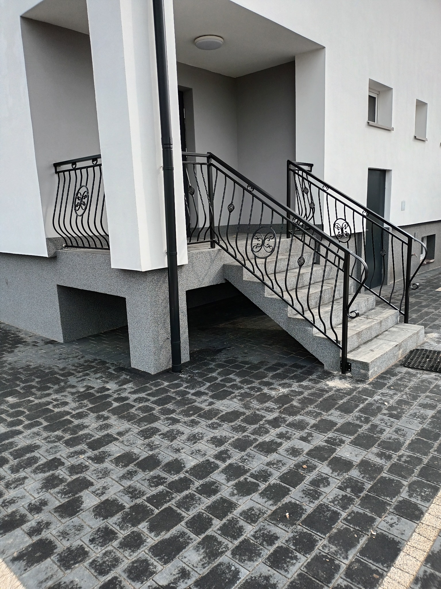 Schody wejściowe do budynku z czarnymi, metalowymi poręczami o zdobieniach, wykończone szarą kostką brukową i jasną elewacją.
