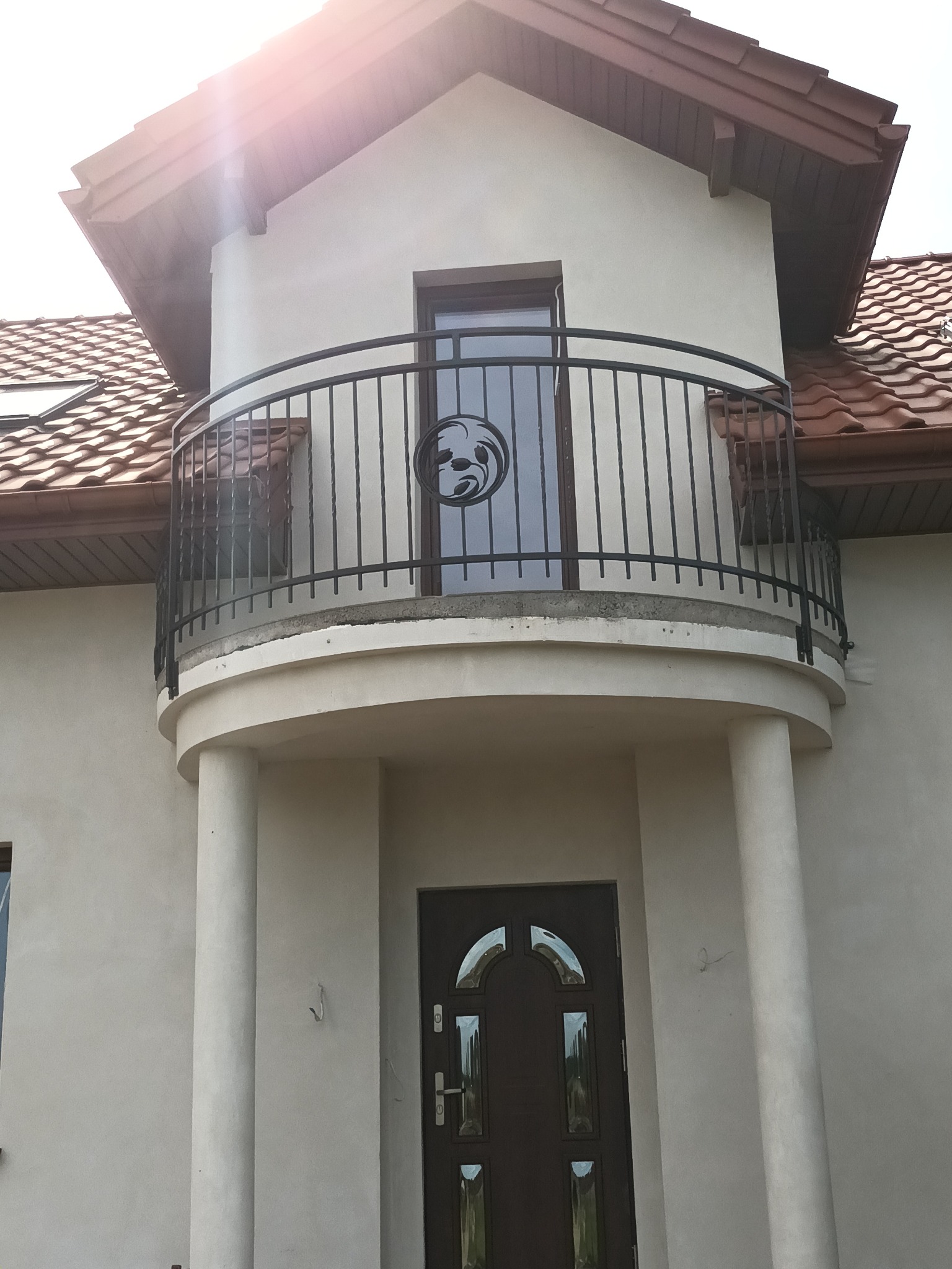 Kuty balkon z czarną, ozdobną barierką z motywem roślinnym w szklanym panelu, na tle jasnej elewacji domu z brązowym dachem.