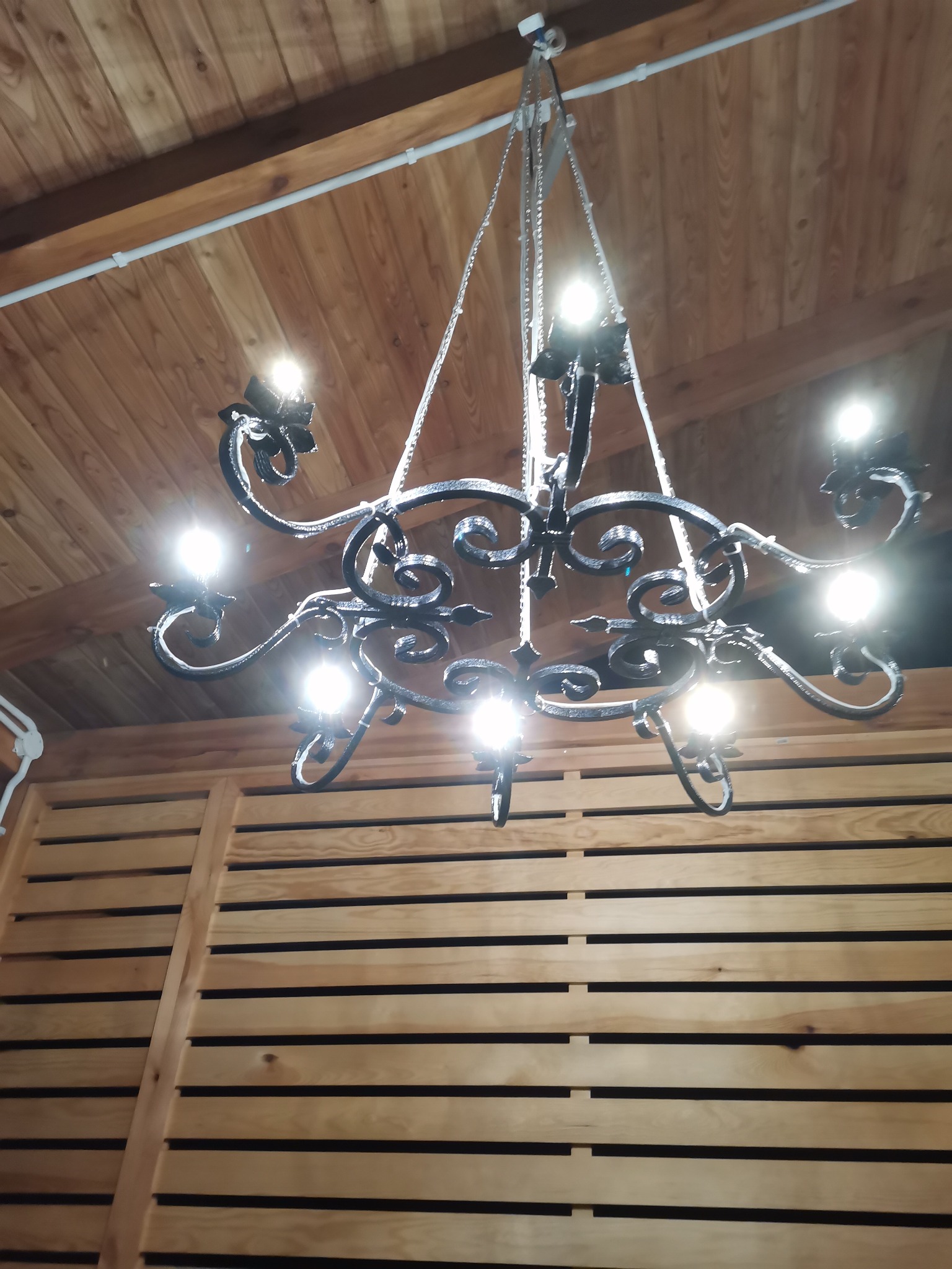 Kuta, metalowa lampa wisząca z żarówkami, zawieszona na tle drewnianego sufitu i ściany z paneli.