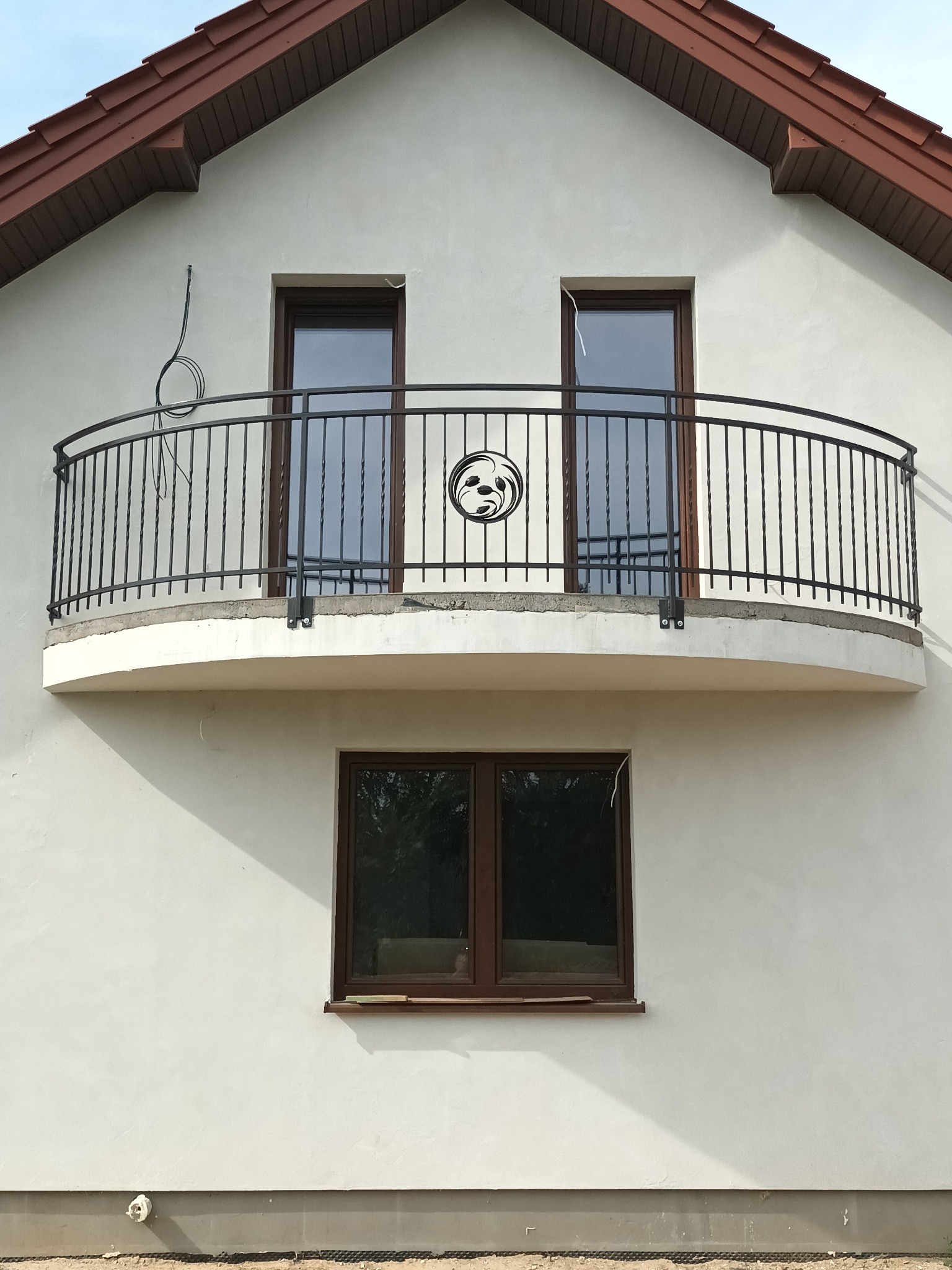Półokrągły balkon z czarną, metalową balustradą z ozdobnym elementem florystycznym na tle białej elewacji domu z brązowym dachem.