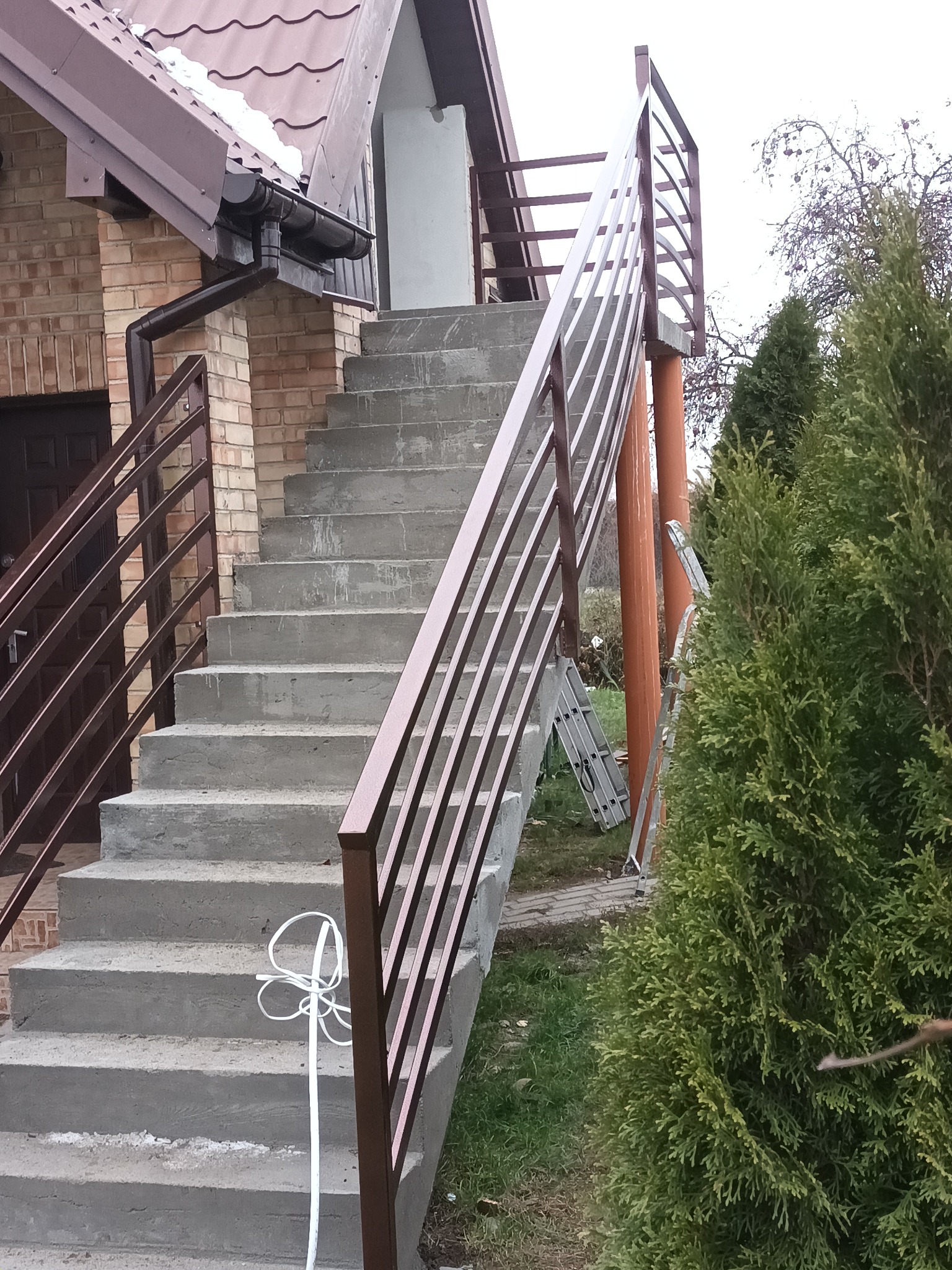 Betonowe schody zewnętrzne z brązową metalową balustradą, widoczne ślady po budowie, w tle drabina i zielony krzew.