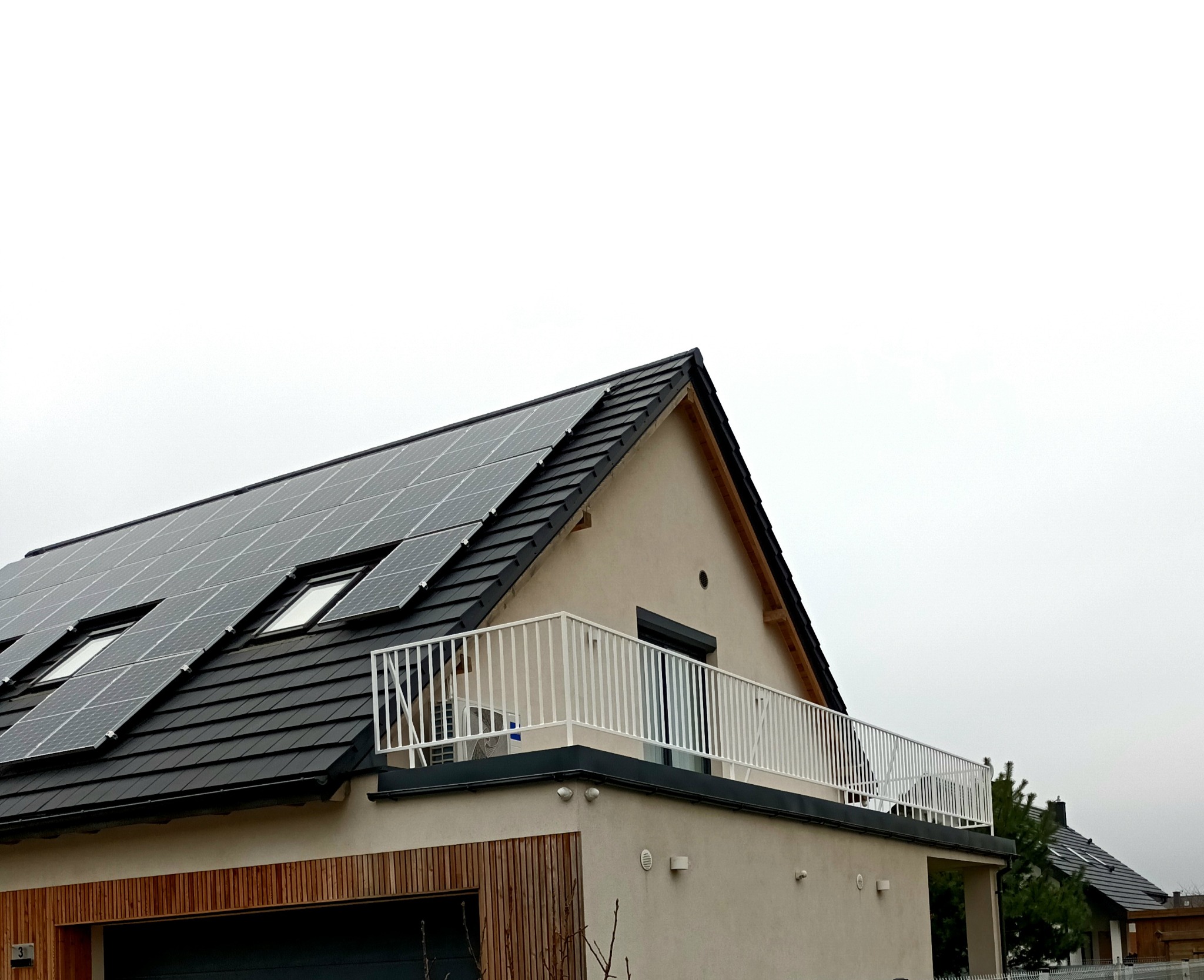 Nowoczesny dom z balkonem zabezpieczonym białą, metalową konstrukcją, panele słoneczne na dachu, widok z dołu na tle pochmurnego nieba.