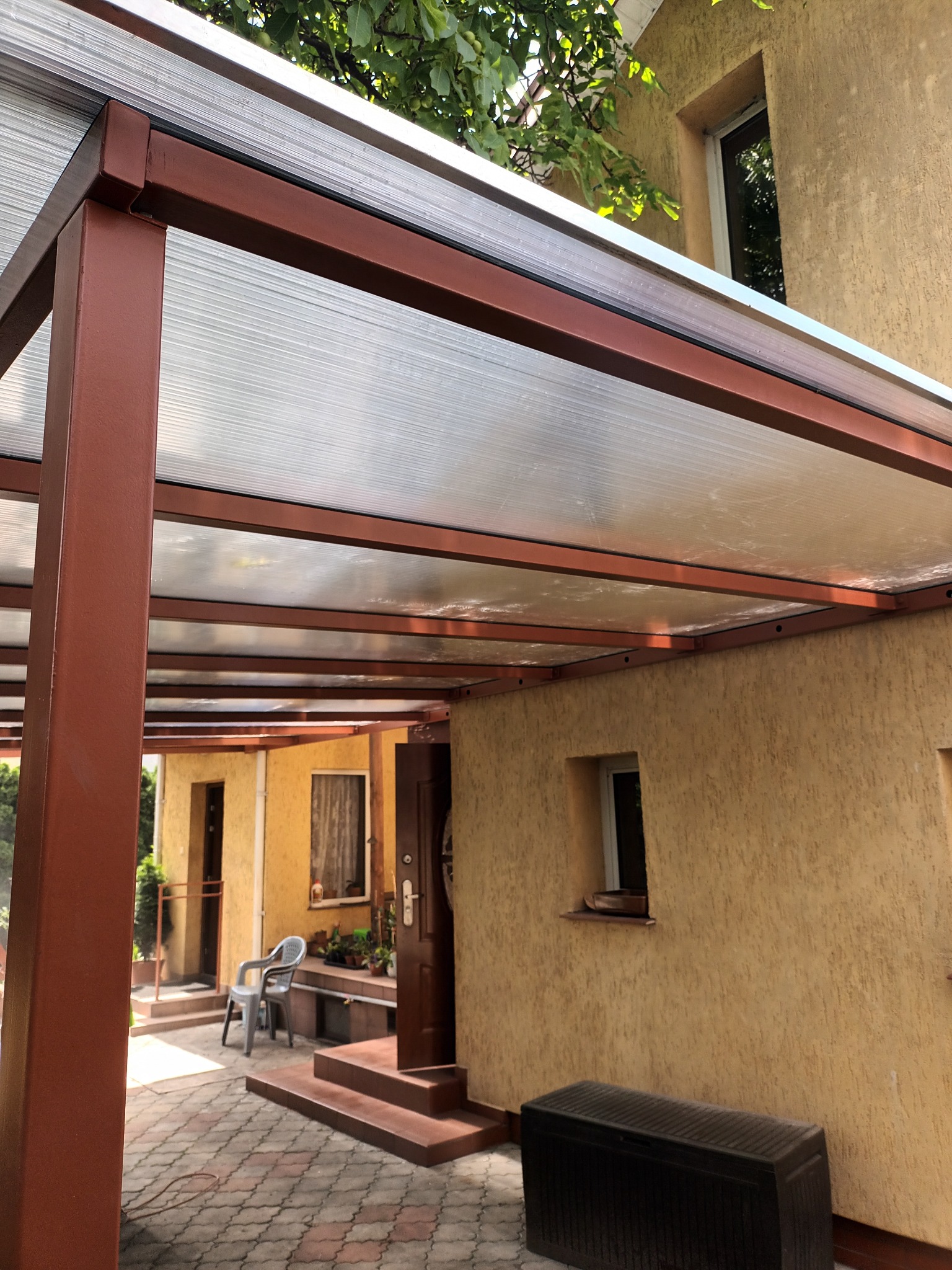 Zadaszenie tarasu z brązowej metalowej konstrukcji i przezroczystego poliwęglanu, widok z dołu na tle elewacji budynku i drzew