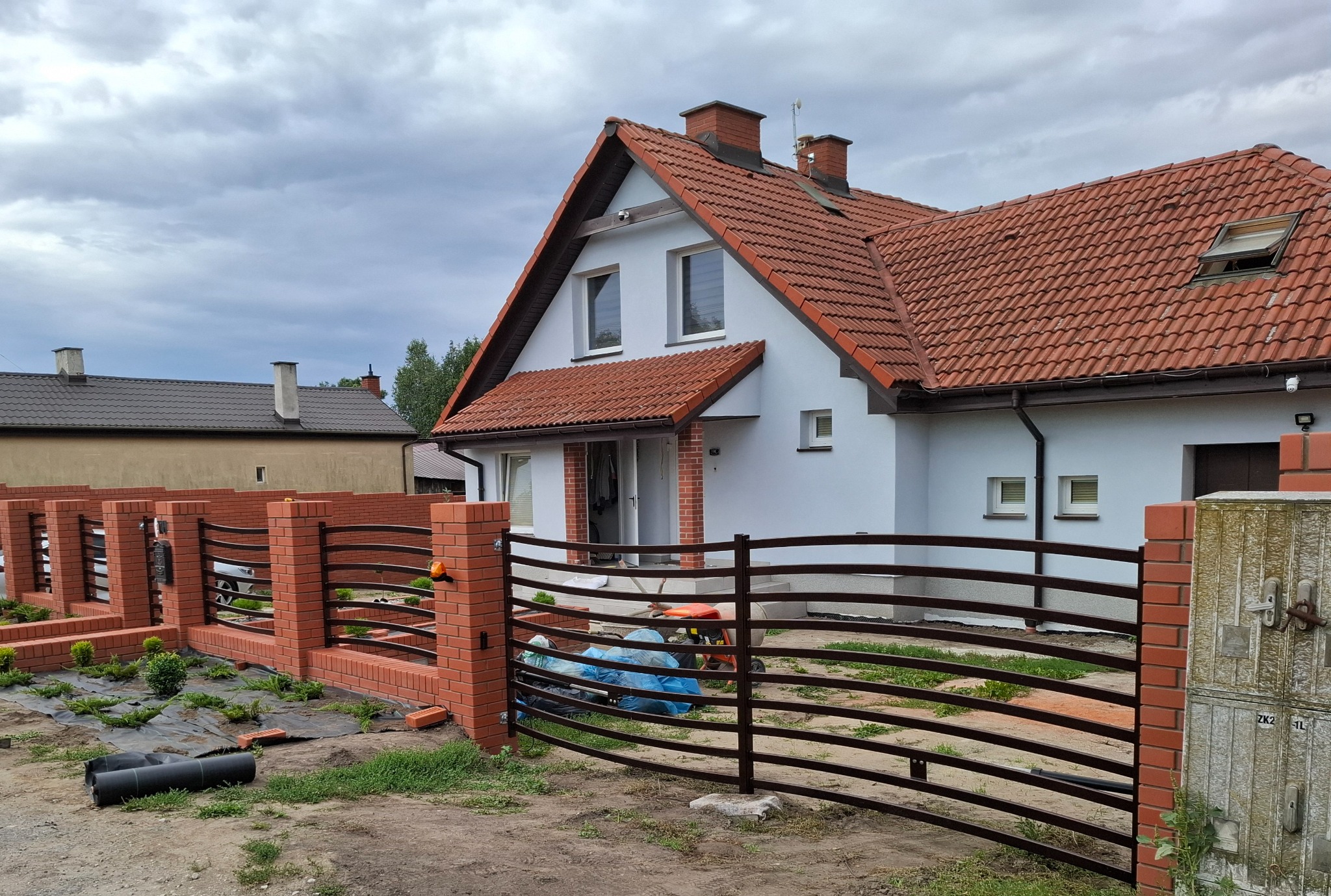 Nowoczesne ogrodzenie z metalowymi przęsłami i ceglanymi słupkami przed domem z czerwoną dachówką, widoczne prace wykończeniowe w postaci folii i narzędzi.