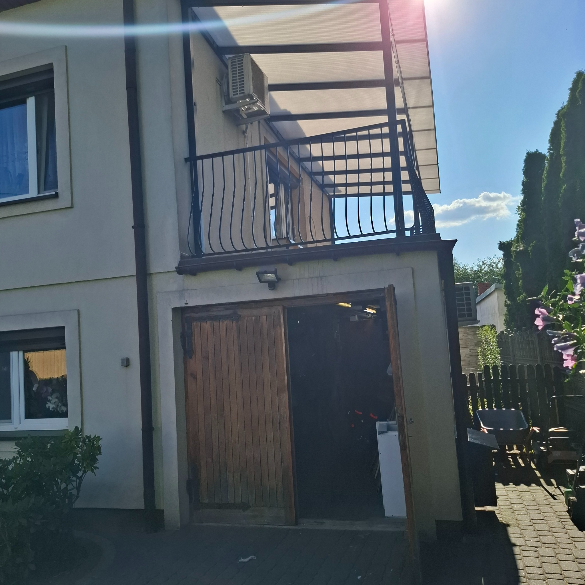 Dwupiętrowy dom z balkonem z czarną, metalową balustradą, zadaszeniem z poliwęglanu i otwartymi drewnianymi drzwiami garażowymi