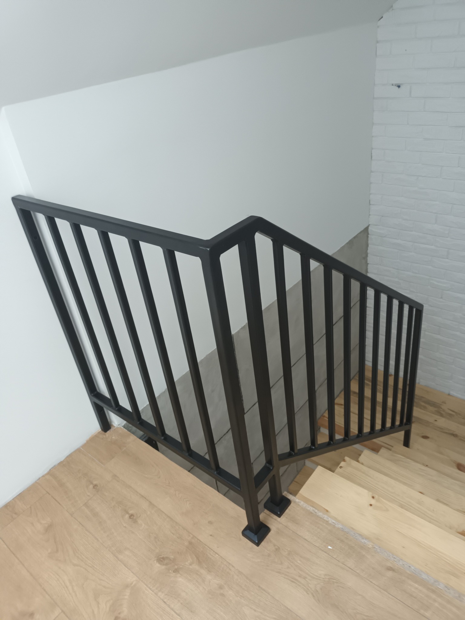 Nowoczesna, czarna, metalowa barierka schodowa z pionowymi prętami, zamontowana na drewnianych schodach w jasnym wnętrzu z elementami białej cegły.
