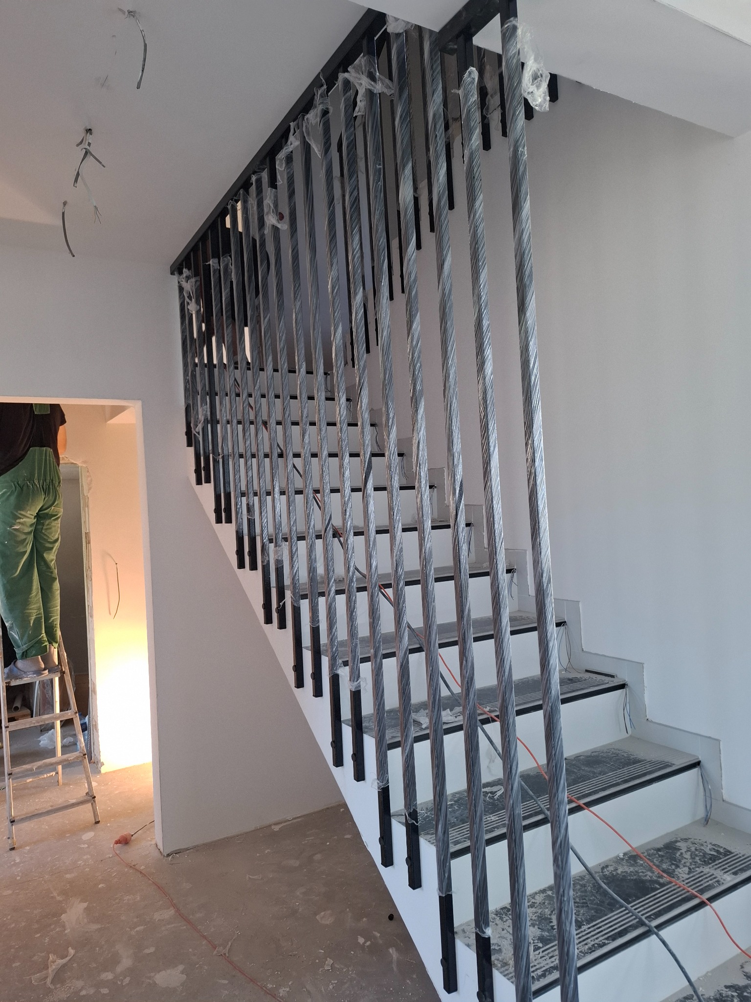 Nowoczesna balustrada schodowa z czarnymi słupkami i srebrnymi, skręconymi prętami, jeszcze w folii ochronnej, w jasnym wnętrzu w trakcie wykończenia.