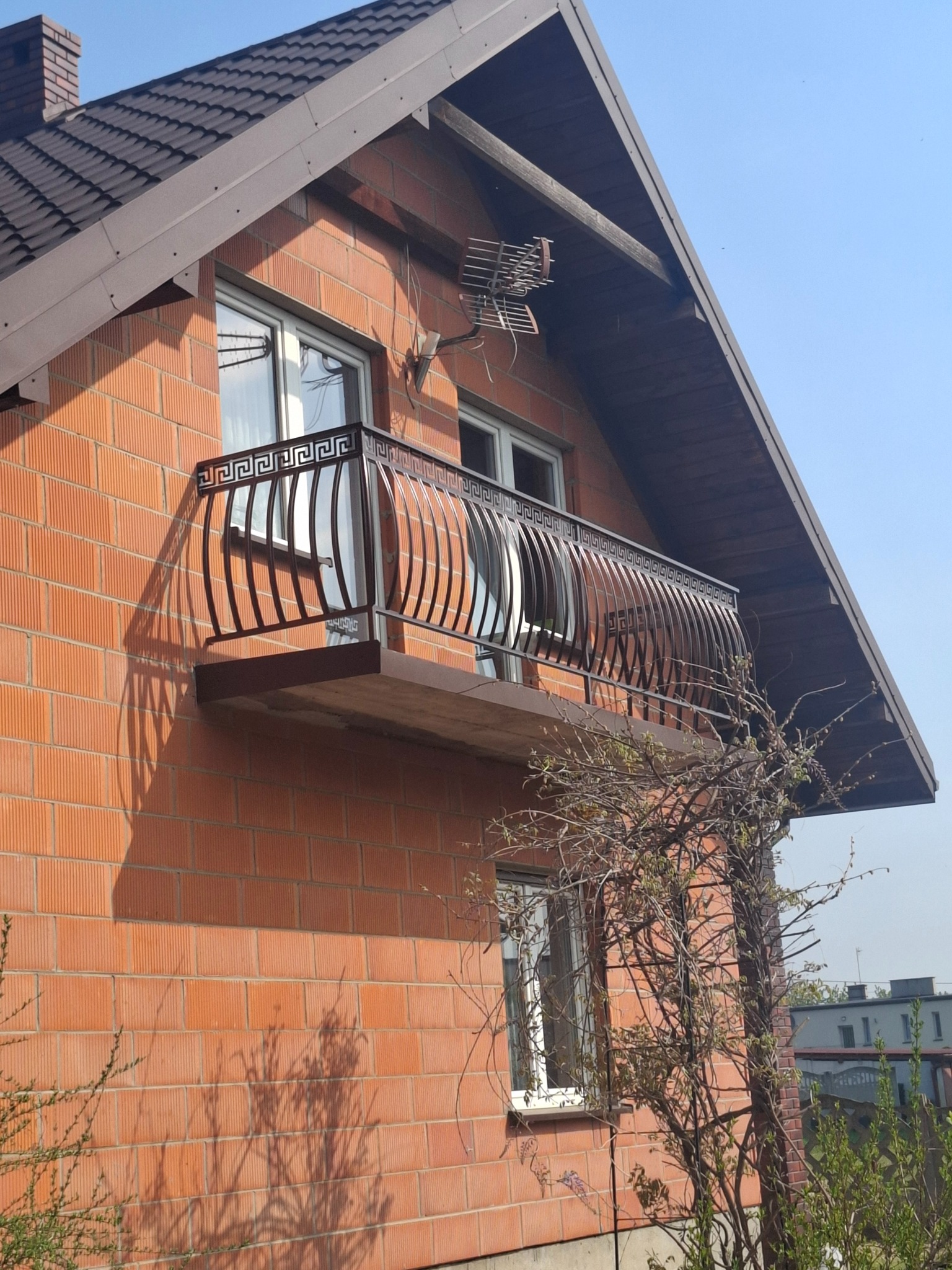 Balkon z metalową balustradą ozdobioną greckim wzorem na tle ceglanej ściany domu z brązowym dachem. Na balkonie widoczne odbicie nieba i drzew.