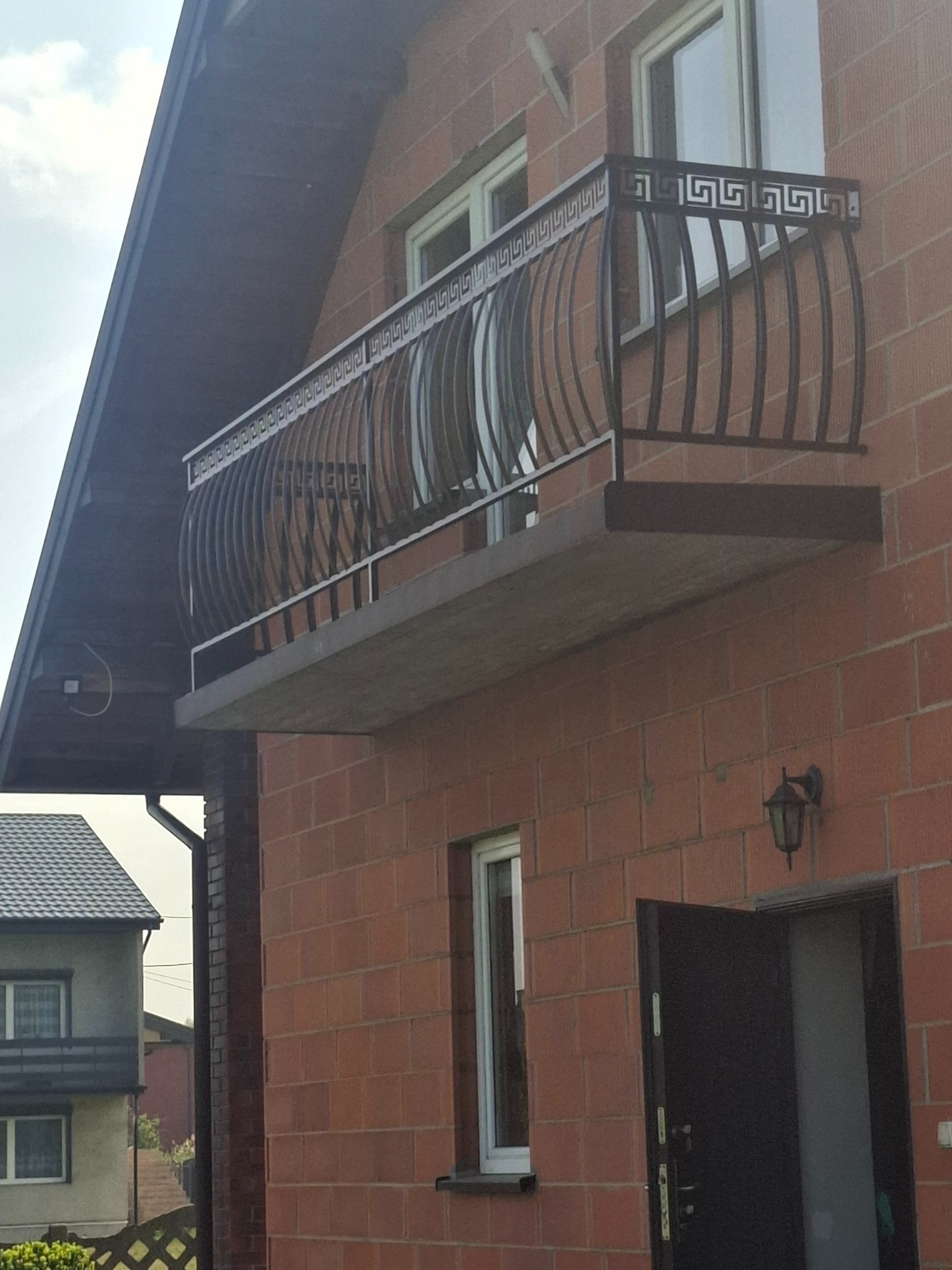 Balkon z dekoracyjną, metalową balustradą w stylu greckim, zamontowaną na ceglanym budynku. Widoczna lampa zewnętrzna i fragment sąsiedniego domu.