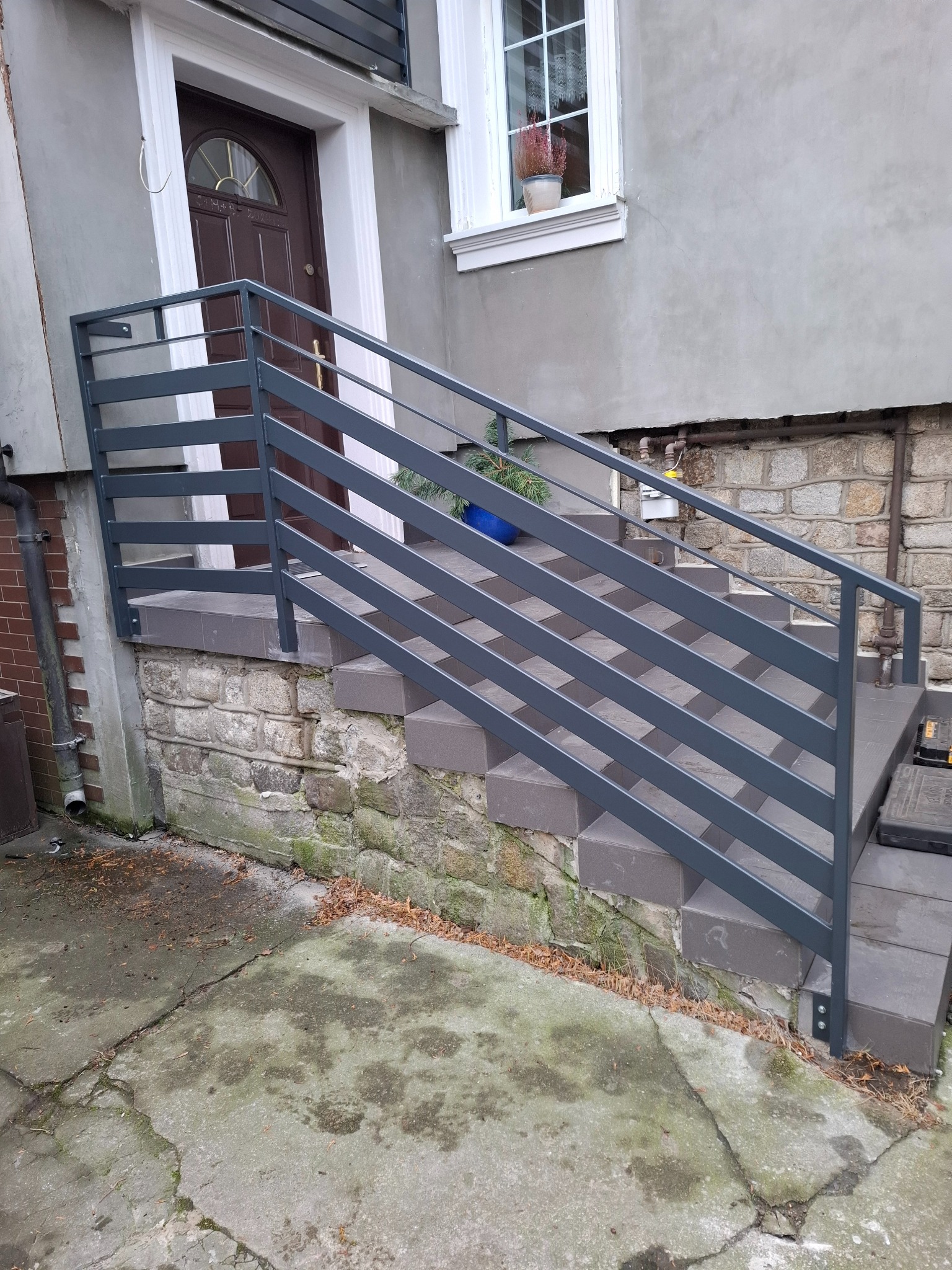 Nowoczesna, szara balustrada metalowa z poziomymi szczeblami, zamontowana przy schodach wejściowych do budynku z kamienną podmurówką i brązowymi drzwiami.