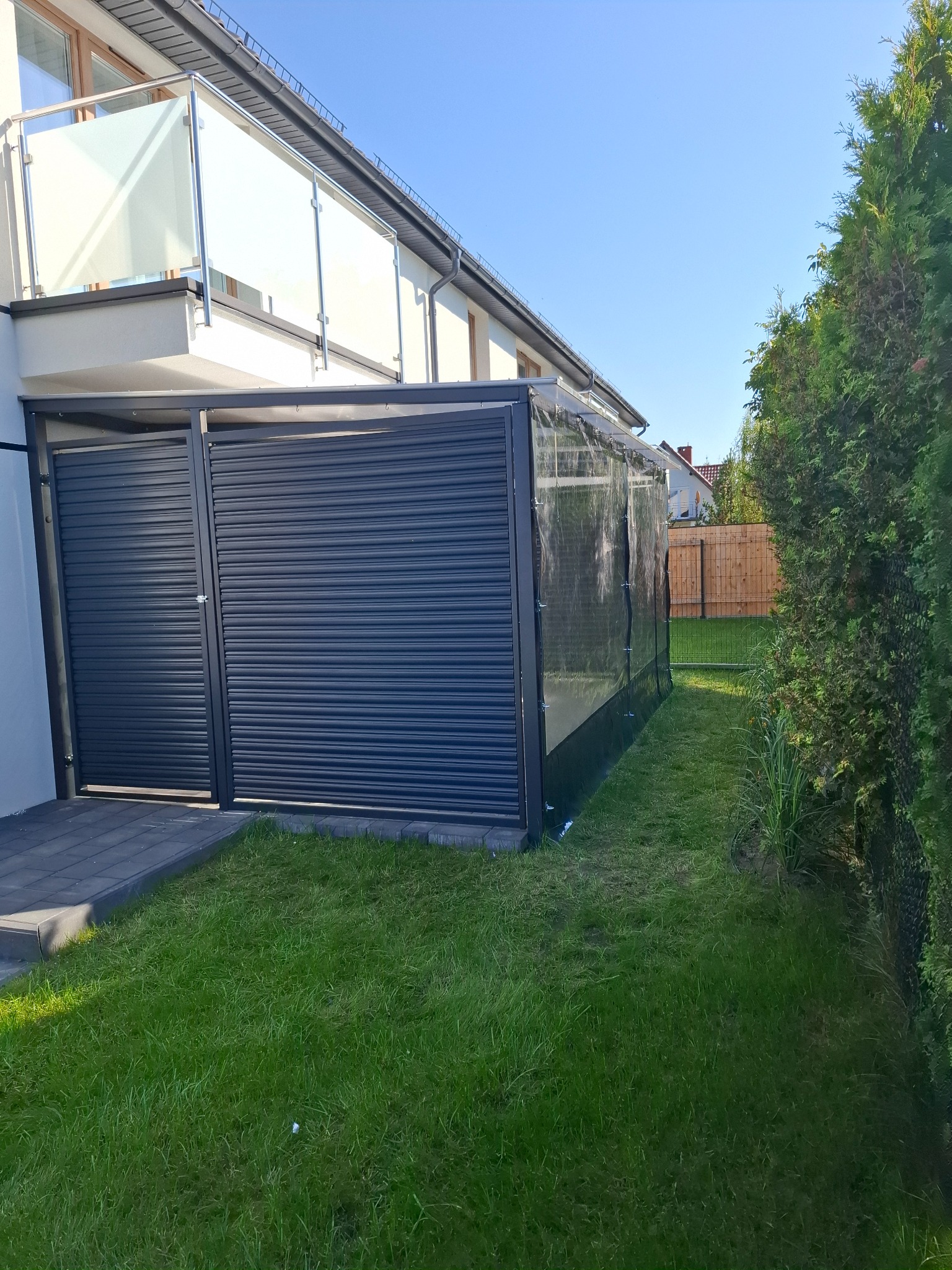 Nowoczesna zabudowa tarasu z roletami i przezroczystymi panelami, przylegająca do domu z balkonem z balustradą ze szkła i stali nierdzewnej, widok z zewnątrz na trawnik.
