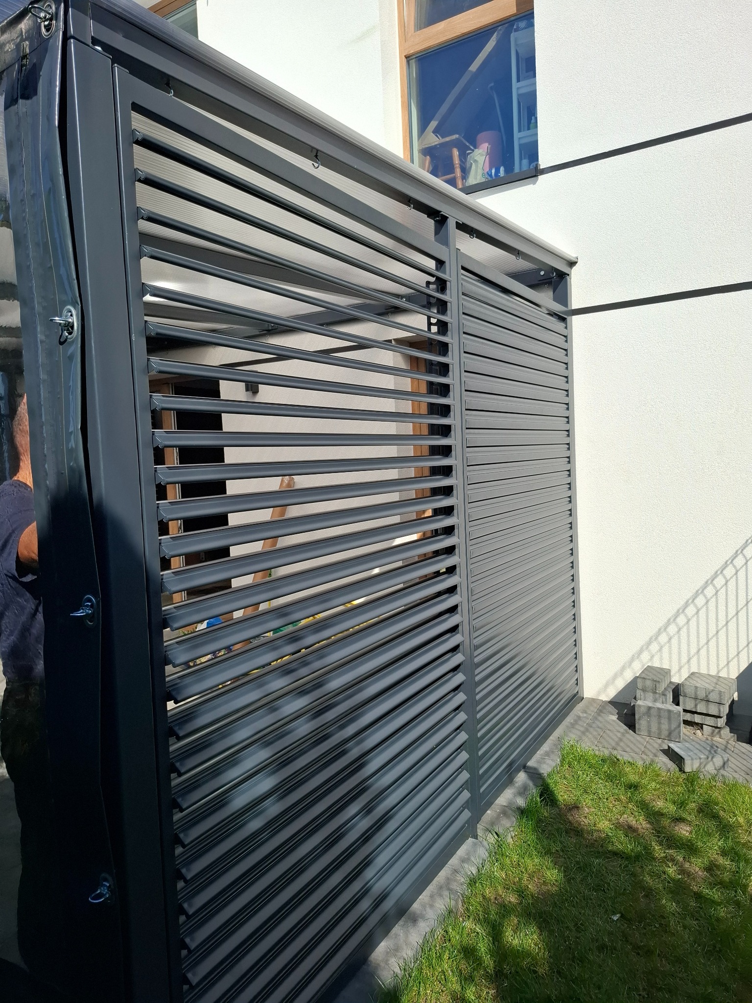 Nowoczesna, szara pergola z lamelami, częściowo zasłonięta folią ochronną, na tle białej ściany budynku i trawnika. Widoczne elementy montażowe.