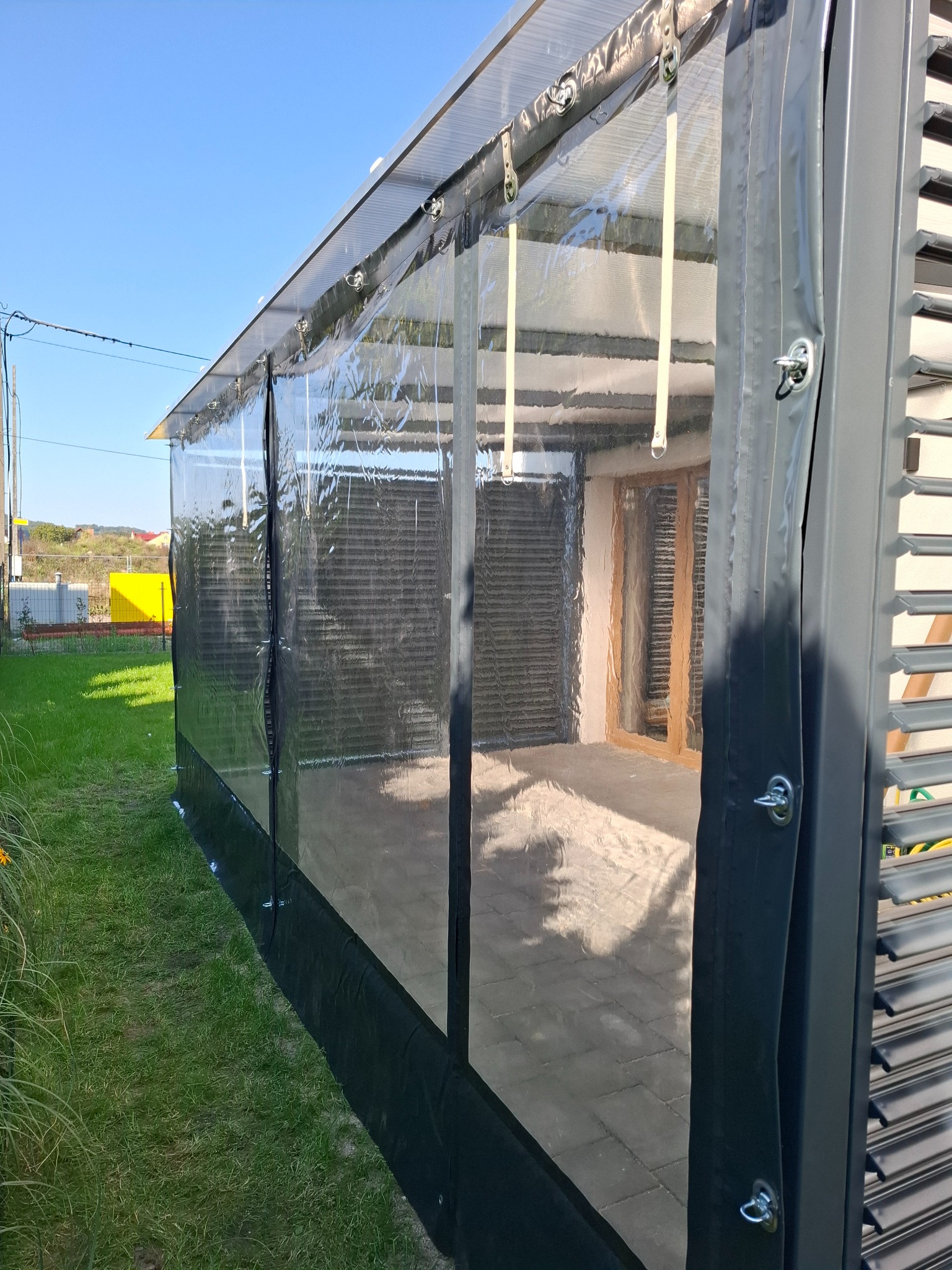 Zadaszenie tarasu z przezroczystą folią i metalową konstrukcją w kolorze grafitowym, widok z zewnątrz na tle trawnika i błękitnego nieba.