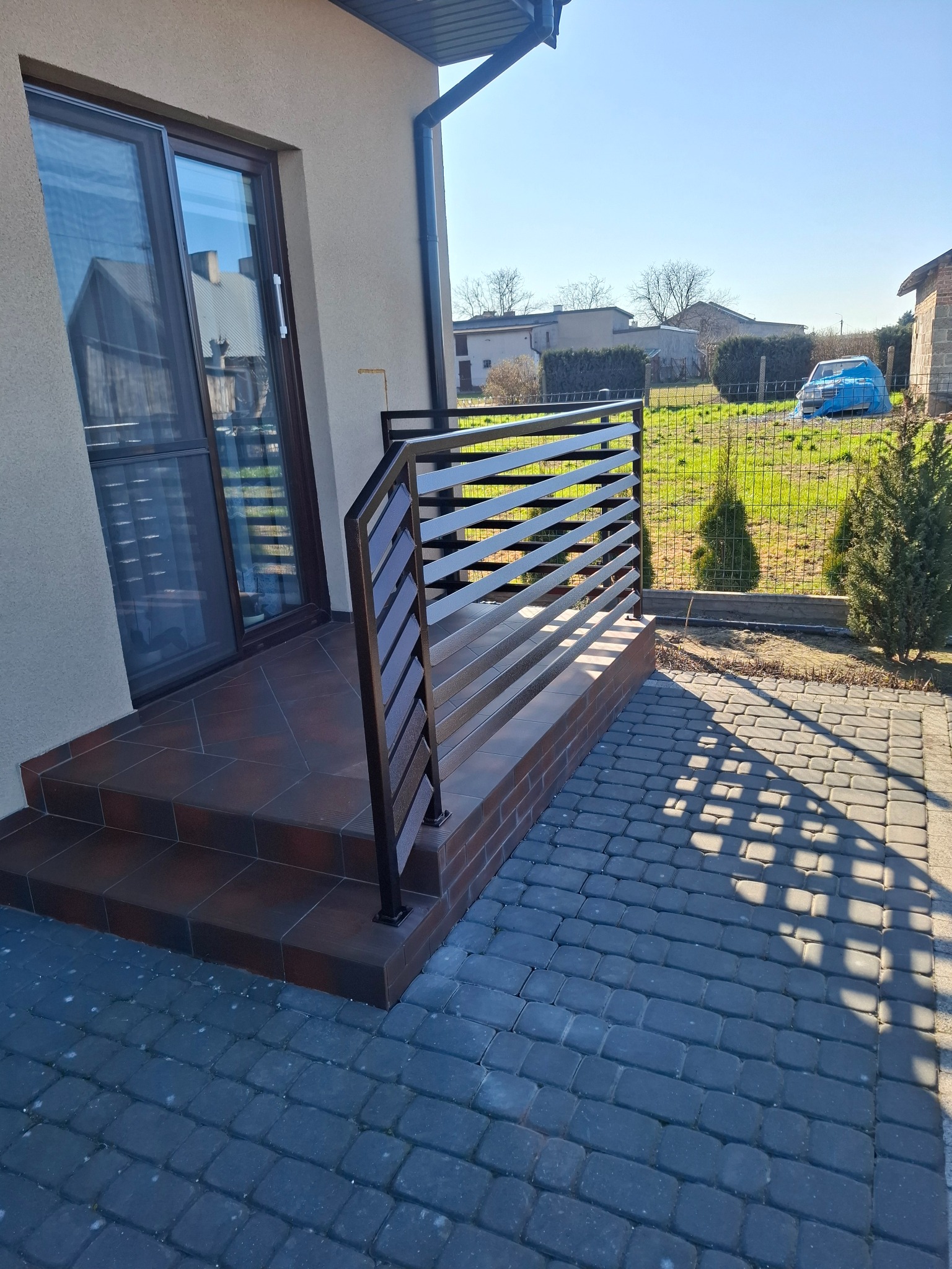 Nowoczesna, pozioma balustrada z ciemnego metalu, zamontowana przy schodach wejściowych do domu. Widoczne płytki na schodach i kostka brukowa na podwórku.