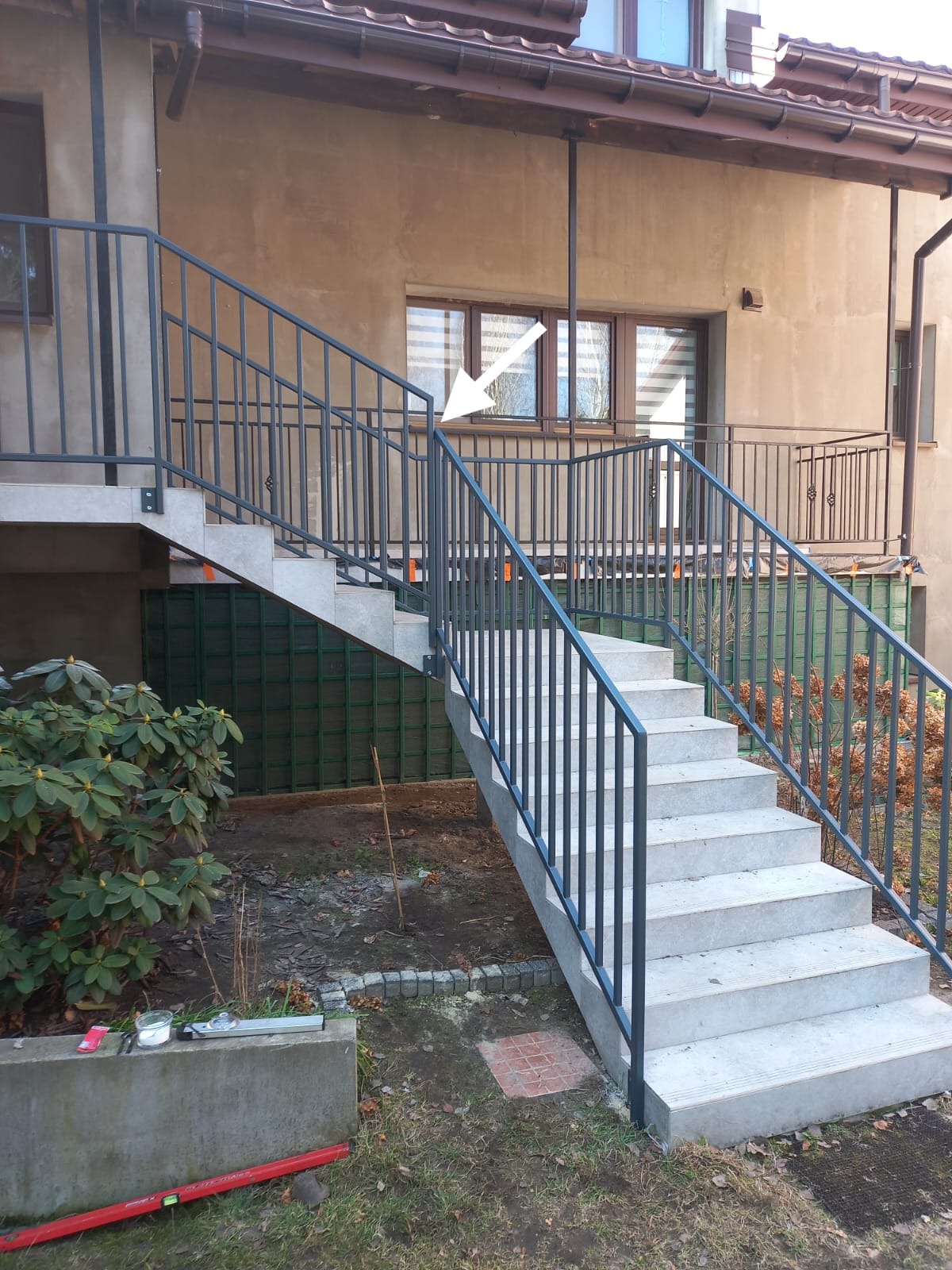 Metalowe poręcze schodowe w kolorze grafitowym, prowadzące do wejścia domu. Widoczne schody z jasnych płyt, zielona roślinność i fragment elewacji budynku.