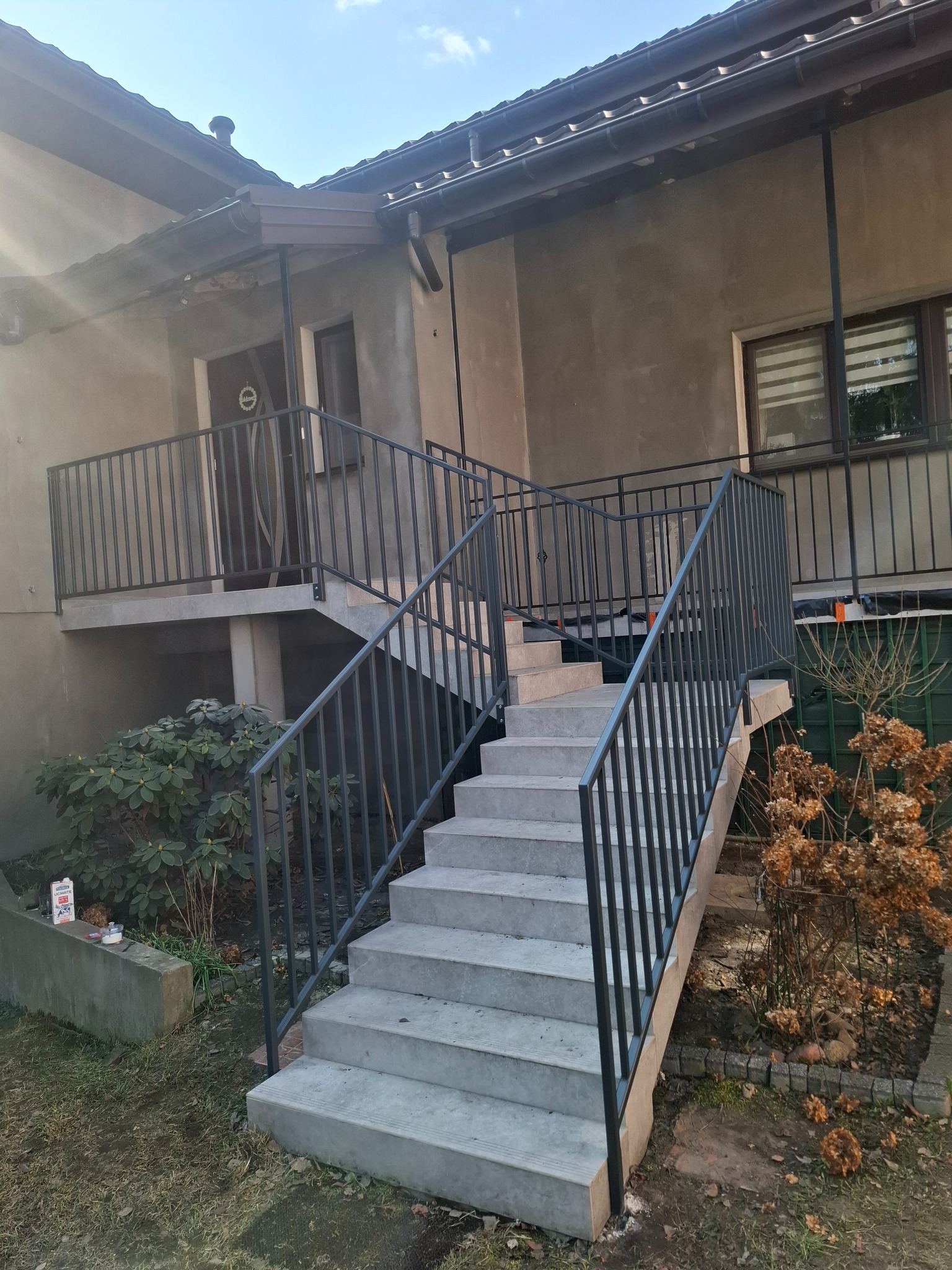 Schody wejściowe z ciemną, metalową barierką, prowadzące do domu z szarą elewacją. Widoczne rośliny i fragment okna z roletami.
