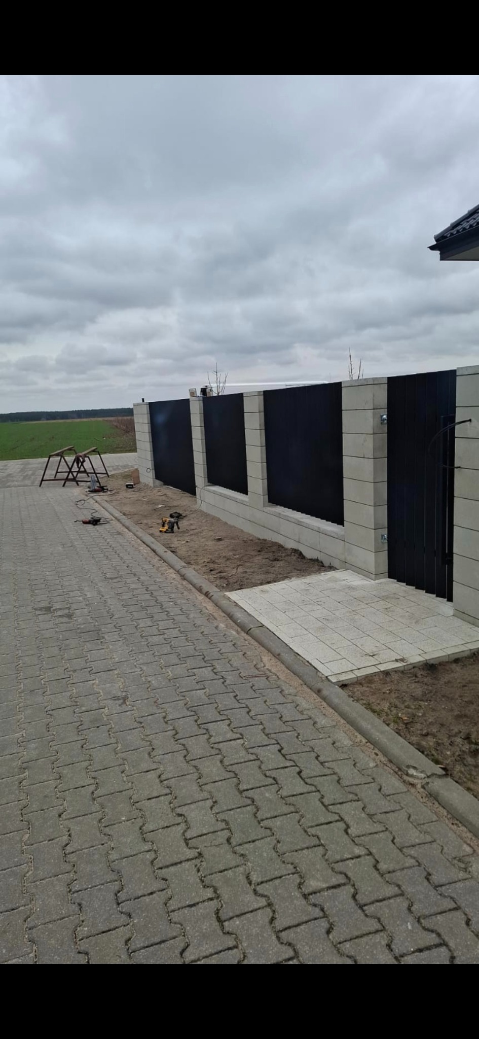 Nowoczesne ogrodzenie z betonowych bloczków i czarnych paneli, furtka z pionowych profili. Widoczne narzędzia i stojaki, sugerujące montaż w toku.