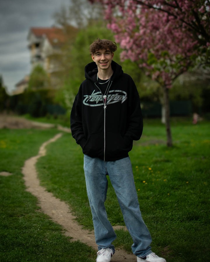 Portret młodego mężczyzny w czarnej bluzie z kapturem i jeansach, stojącego na ścieżce w parku z kwitnącym drzewem wiśni w tle.