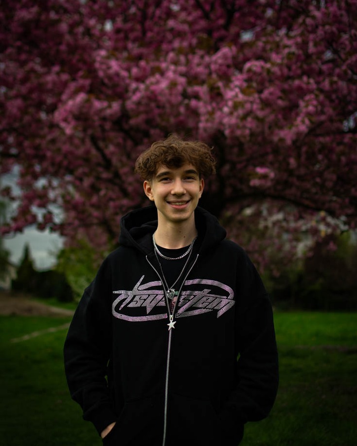Portret uśmiechniętego nastolatka w czarnej bluzie z kapturem, na tle kwitnącego drzewa wiśni o różowych kwiatach.