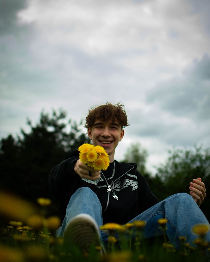 Uśmiechnięty młody mężczyzna siedzi na łące pełnej mleczy, trzymając bukiet tych kwiatów w wyciągniętej dłoni, ubrany w czarną bluzę z białym wzorem i srebrny łańcuszek.