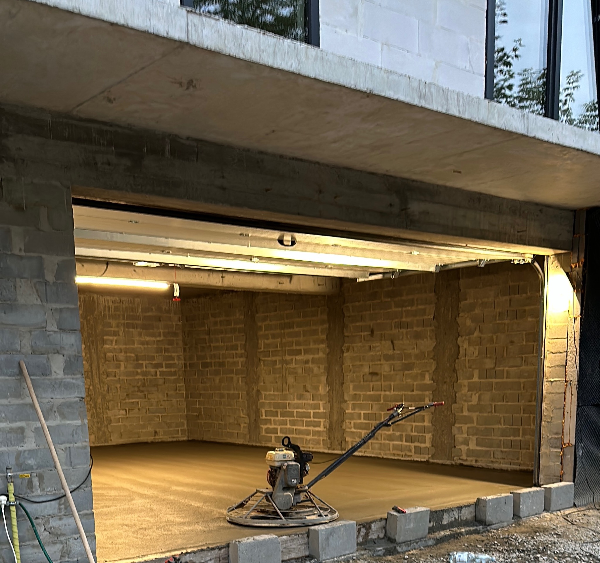 Zacieraczka mechaniczna na świeżo wylanej, jasnej posadzce w surowym garażu z ceglanymi ścianami i widocznym oświetleniem.