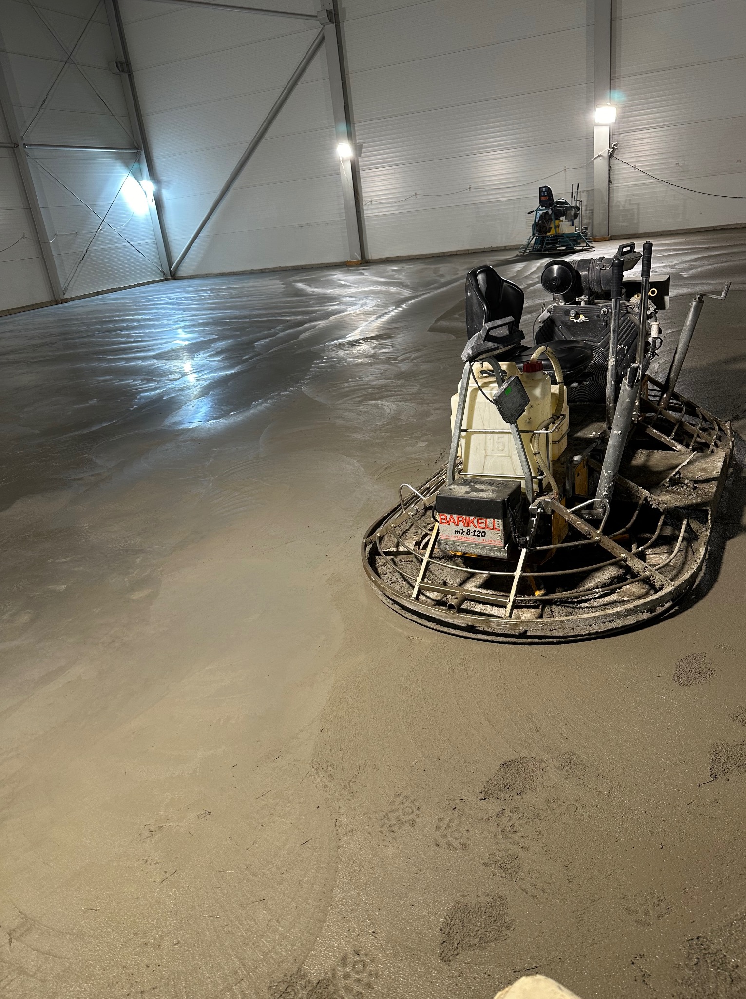 Zacieraczka mechaniczna Barikell mk8-120 w trakcie wygładzania wylewki betonowej posadzki przemysłowej w hali, widoczne ślady butów na świeżym betonie i druga maszyna w tle.