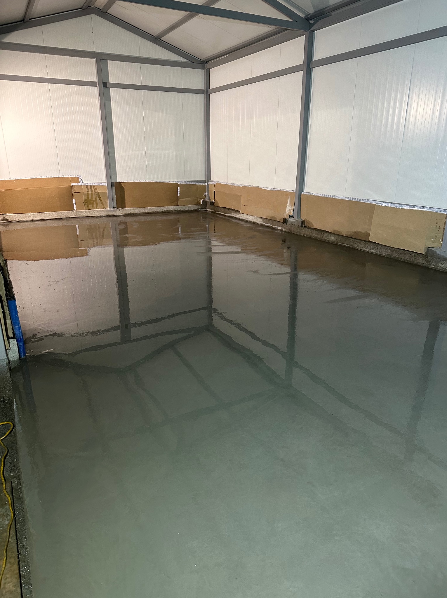 Świeżo wylana, mokra, szara posadzka w hali przemysłowej z widocznymi kartonami zabezpieczającymi ściany i metalową konstrukcją dachu.
