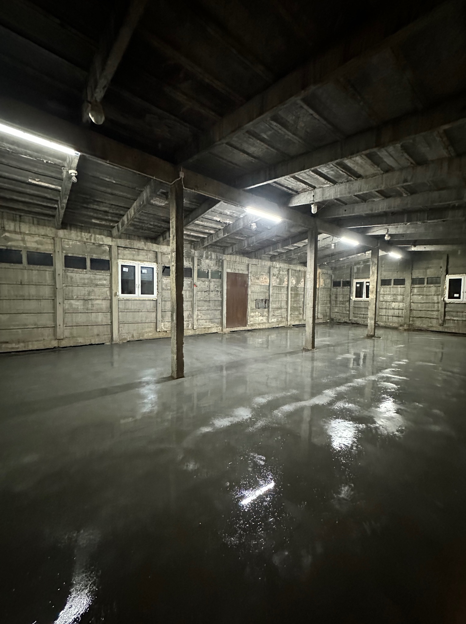 Świeżo wylana, mokra, szara posadzka w hali przemysłowej z betonowymi ścianami i drewnianym stropem, oświetlona lampami jarzeniowymi.