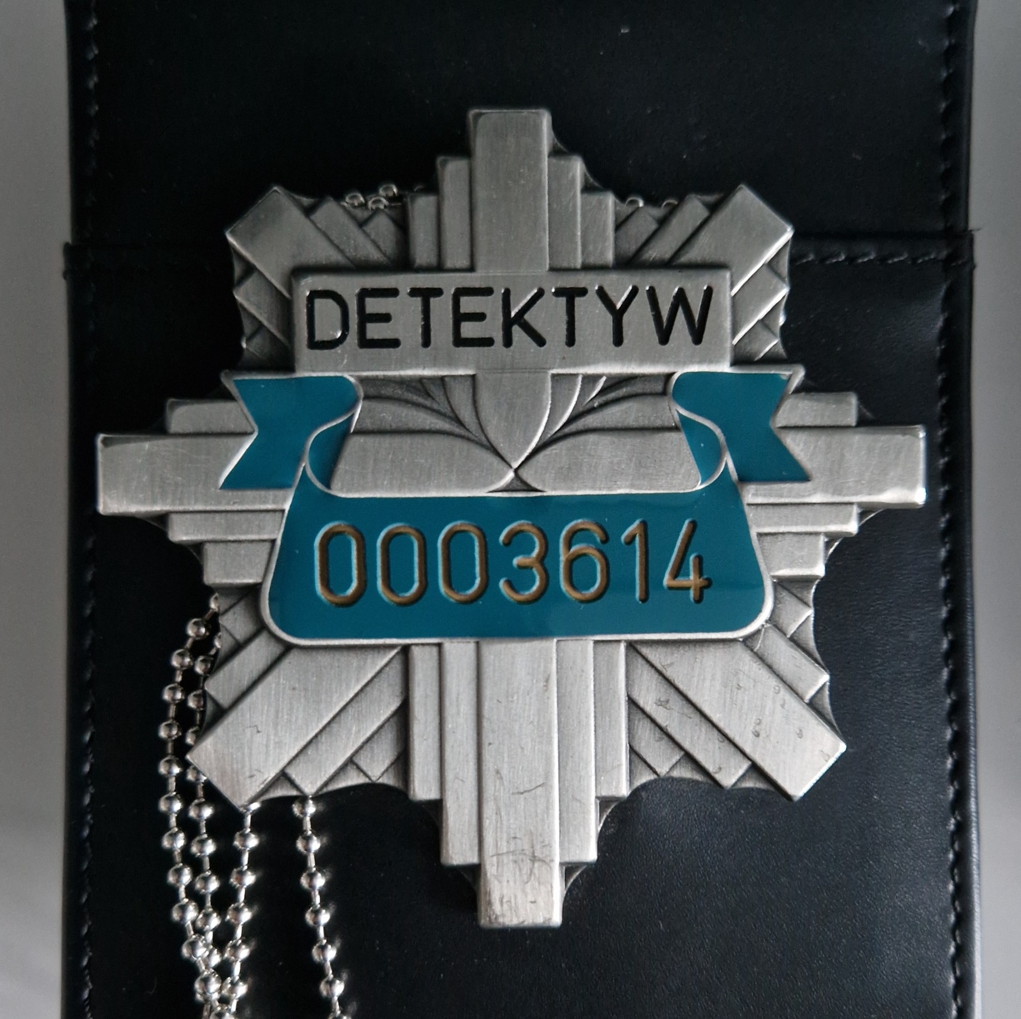 Metalowa odznaka z napisem 'Detektyw' i numerem 0003614, przypięta do czarnego skórzanego etui.