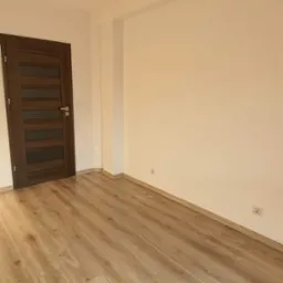 Jasne panele podłogowe z wyraźnym rysunkiem drewna w wyremontowanym pokoju z brązowymi drzwiami i beżową lampą wiszącą.