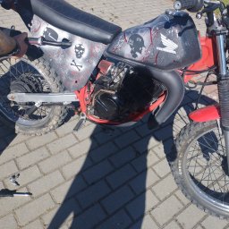 Zrób to sam - Customowy motocykl Honda z czarną tapicerką z czerwonym przeszyciem, ozdobiony motywami czaszek i plamami farby, stojący na brukowanej nawierzchni obok narzędzi.