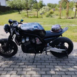 Motocykl typu cafe racer z niestandardowym malowaniem czaszek na baku i kasku, zaparkowany na brukowanej ścieżce w ogrodzie z dekoracyjnymi łukami.