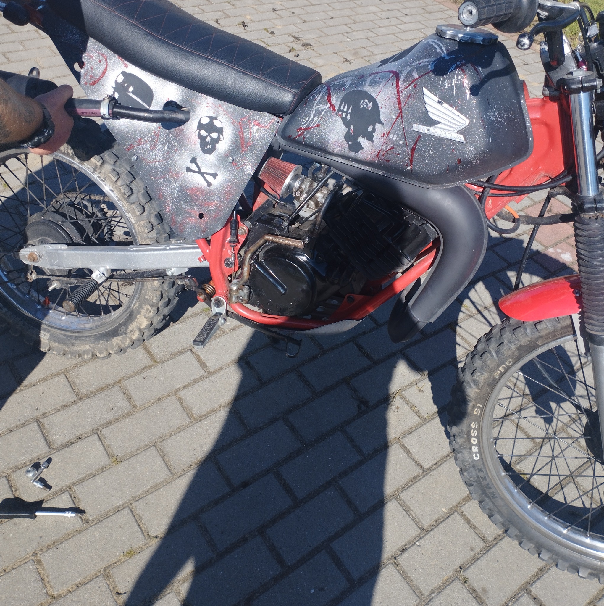 Customowy motocykl Honda z czarną tapicerką z czerwonym przeszyciem, ozdobiony motywami czaszek i plamami farby, stojący na brukowanej nawierzchni obok narzędzi.