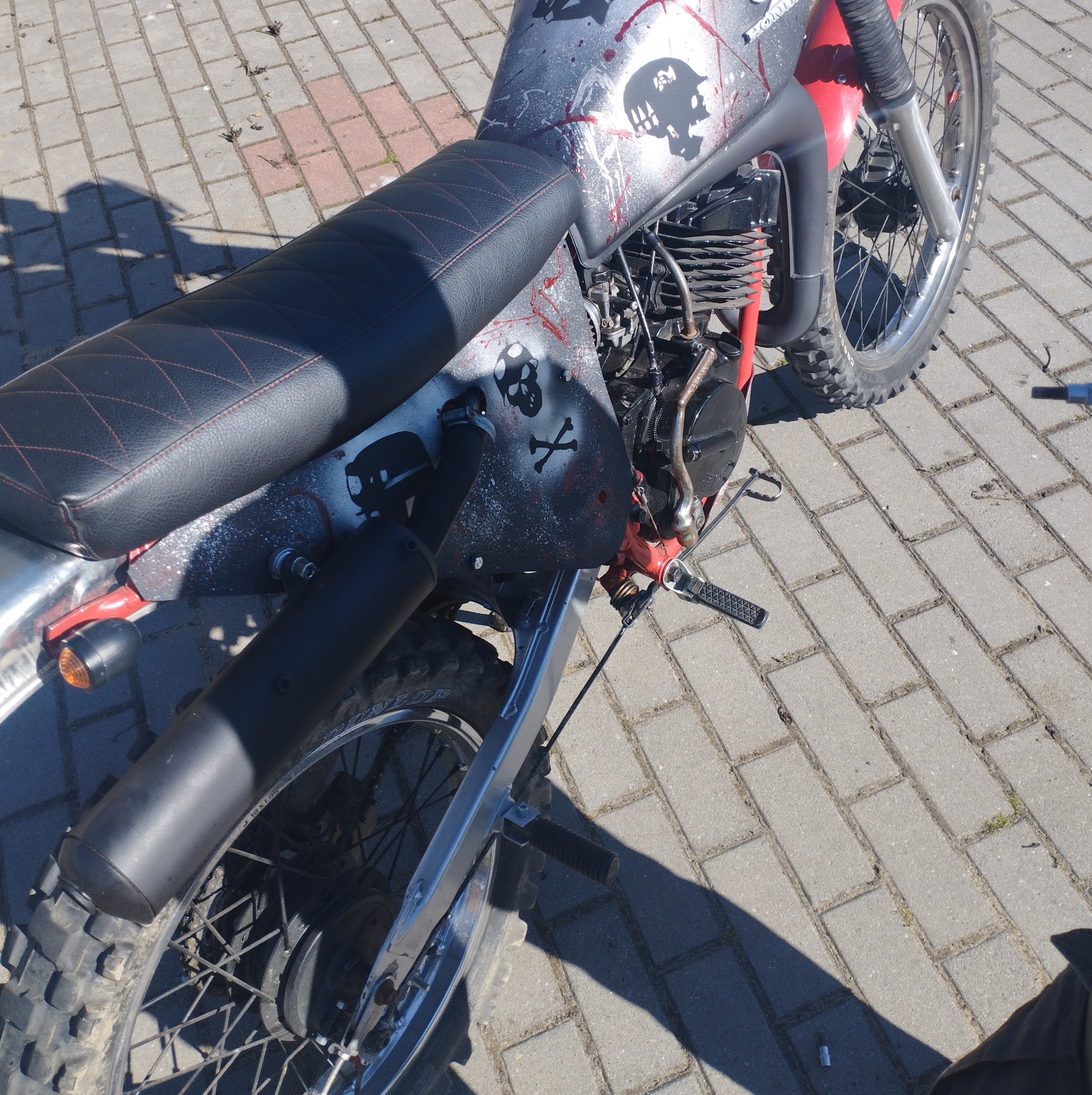 Motocykl crossowy z niestandardowym malowaniem: czaszki, krew i motywy militarne na szarym tle, czarne siedzenie z czerwonymi przeszyciami, widoczne opony terenowe i silnik.