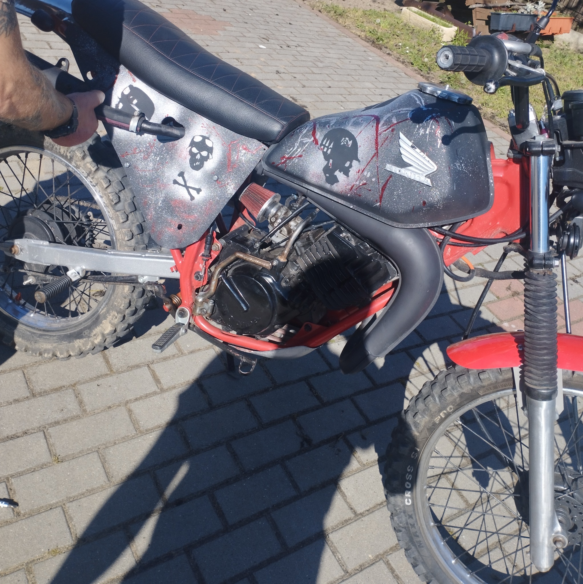 Motocykl Honda z customowym malowaniem czaszek i czerwonych plam, stojący na brukowanym podłożu, widoczne opony terenowe i czerwona rama.