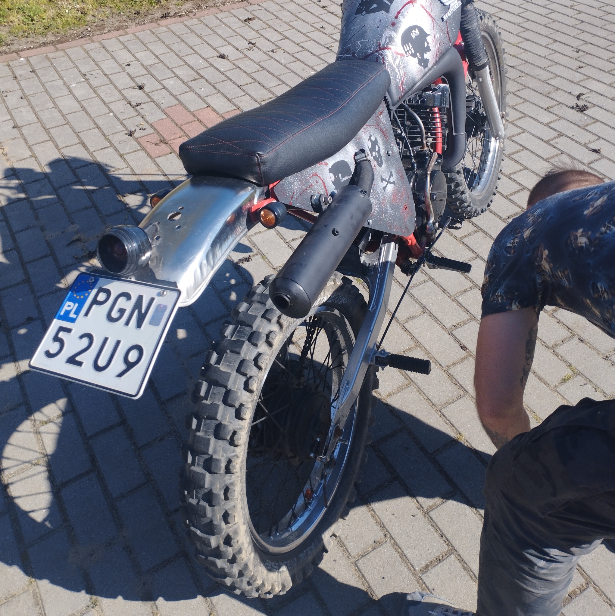 Zmodyfikowany motocykl typu cross z niestandardowym malowaniem czaszek i numerem rejestracyjnym PGN 52U9, stojący na chodniku z kostki brukowej, widziany z tyłu pod kątem, z fragmentem postaci...