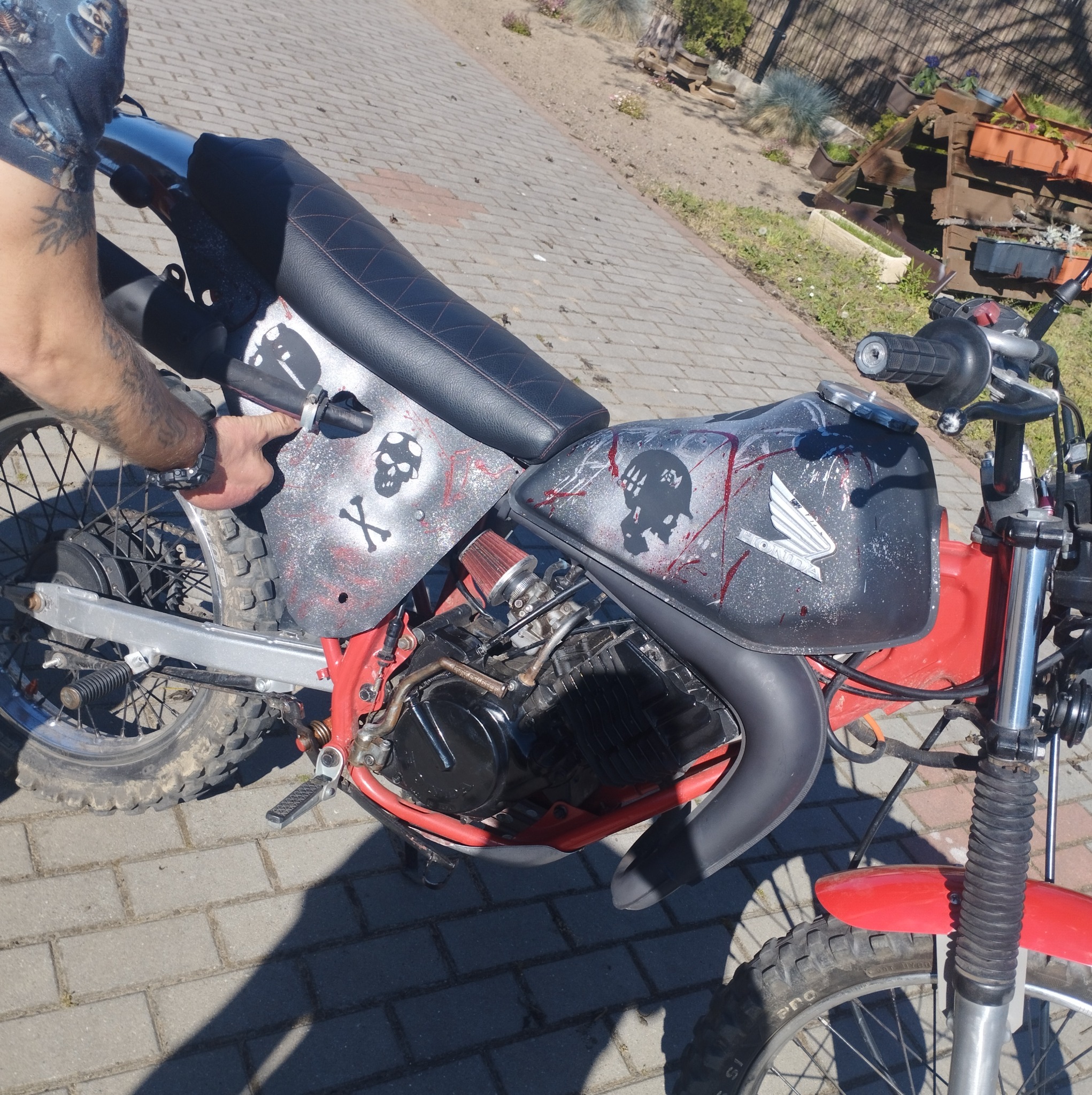 Mężczyzna wskazujący na customowy motocykl Honda z motywem czaszek i czerwonymi akcentami, stojący na brukowanym podwórku z widocznymi elementami ogrodu w tle.