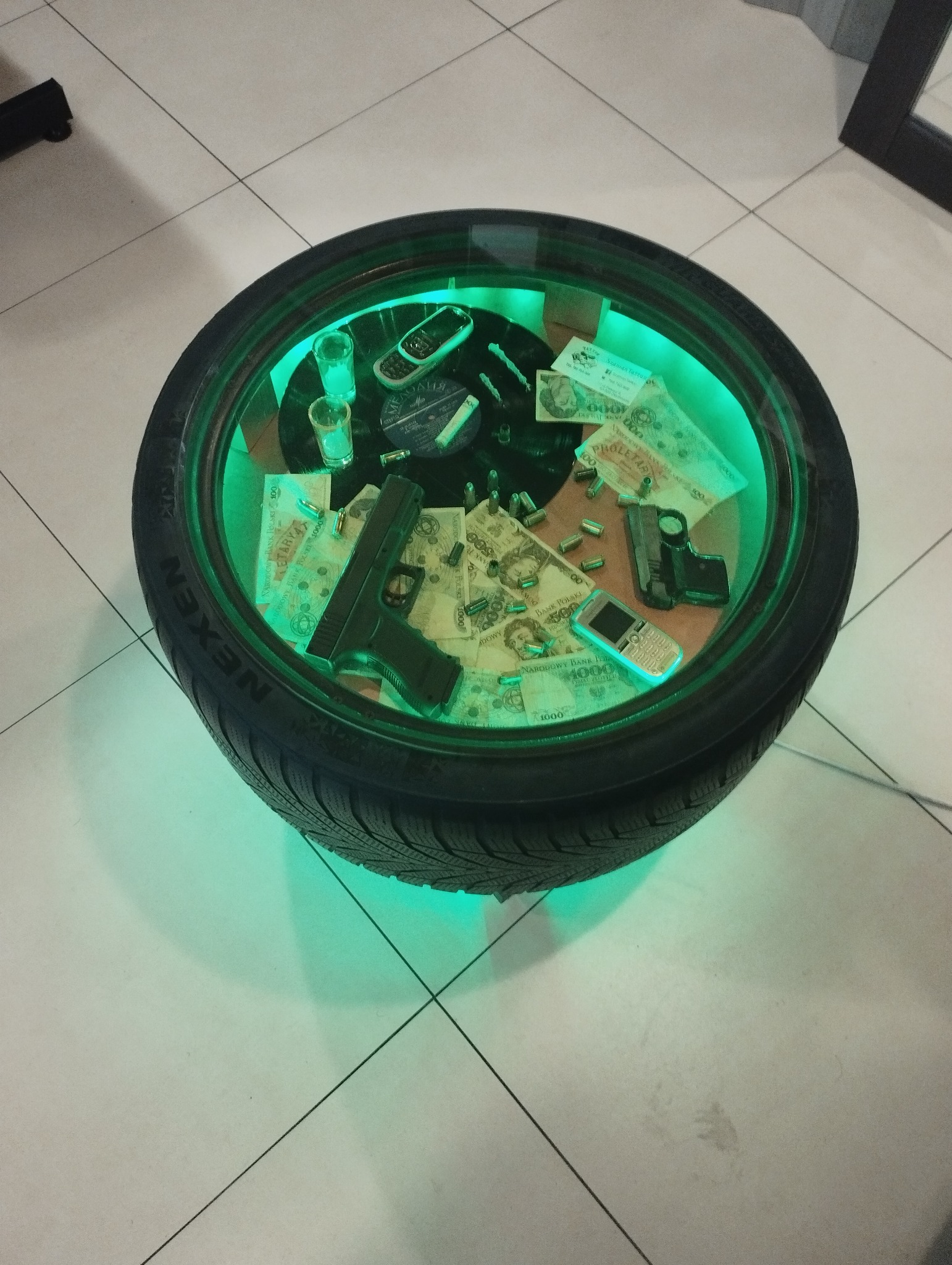 Nietypowy stolik kawowy wykonany z opony samochodowej, podświetlony zielonym światłem, z szybą na górze, na której leżą banknoty, atrapy pistoletów, naboje, stary telefon komórkowy i płyty winylowe.