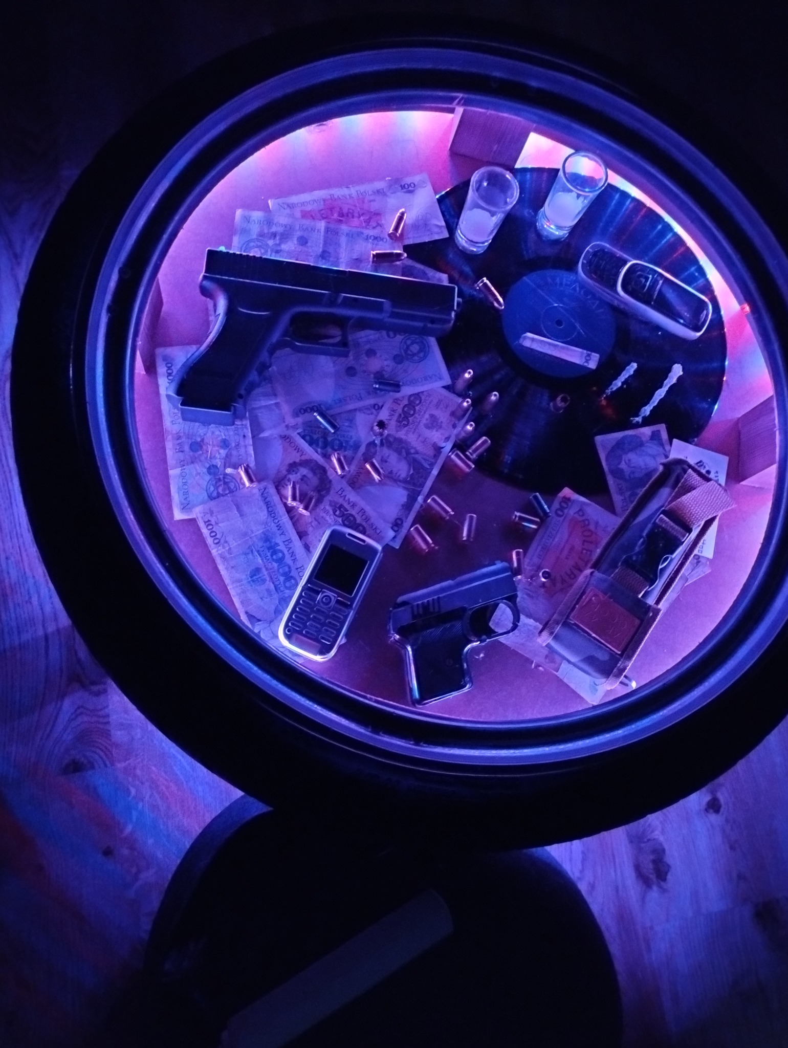 Stolik kawowy wykonany z opony samochodowej, podświetlony na fioletowo, z wnętrzem wypełnionym banknotami, atrapami broni, nabojami, dwoma kieliszkami i telefonem komórkowym, całość ułożona...
