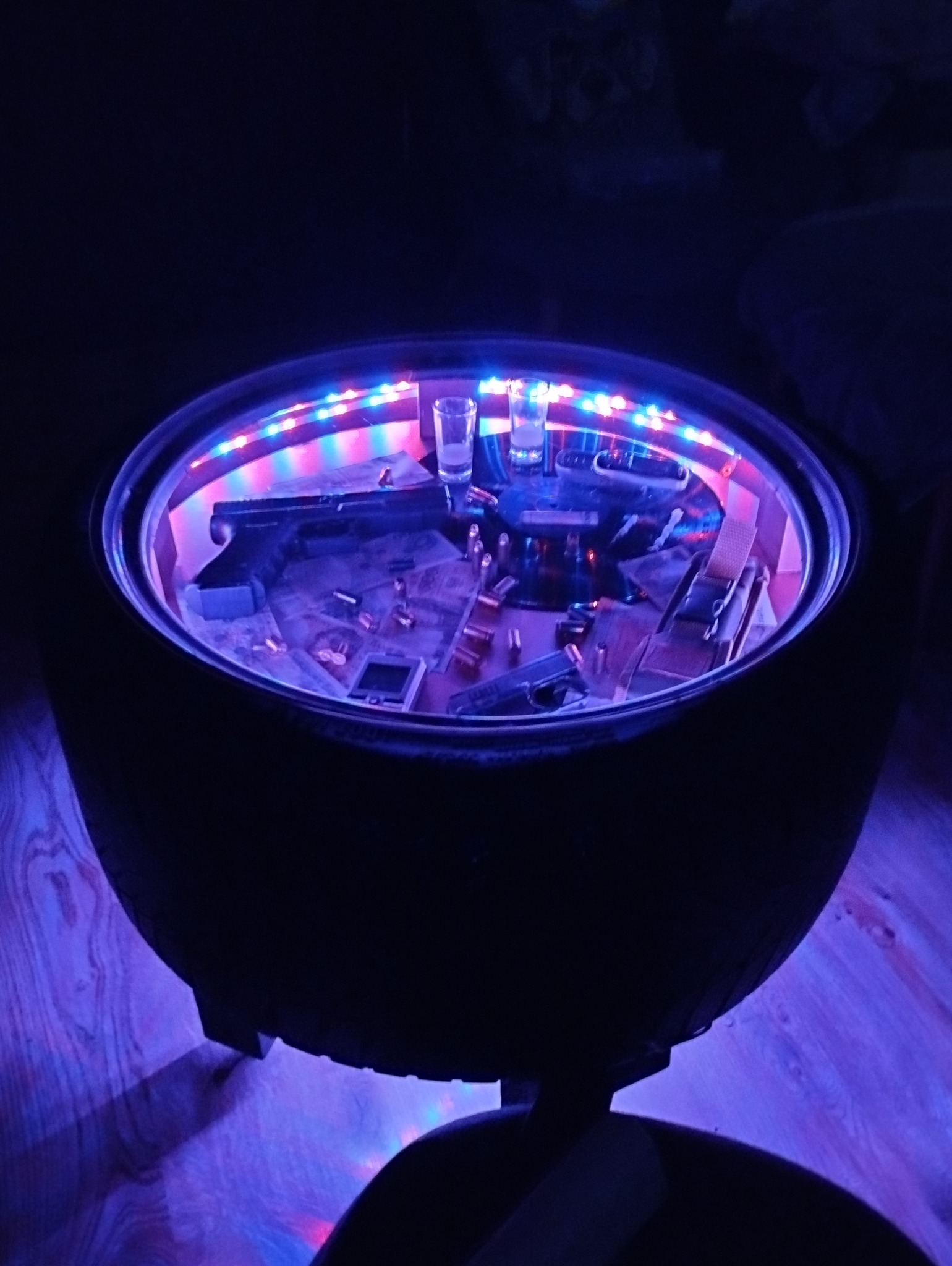 Nietypowy stolik kawowy wykonany ze starej opony, podświetlony kolorowymi diodami LED, z ekspozycją przedmiotów kolekcjonerskich wewnątrz, takich jak repliki broni i łuski nabojów, oraz dwa kieliszki.