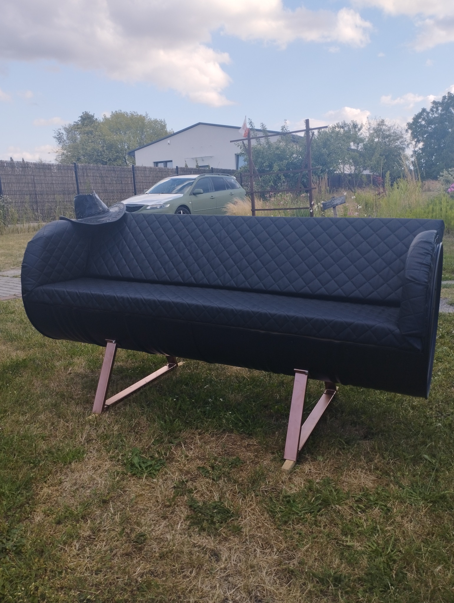 Nietypowa czarna sofa w kształcie walca z pikowanym obiciem, ustawiona na metalowych nogach w ogrodzie, widok na dom i samochód w tle.