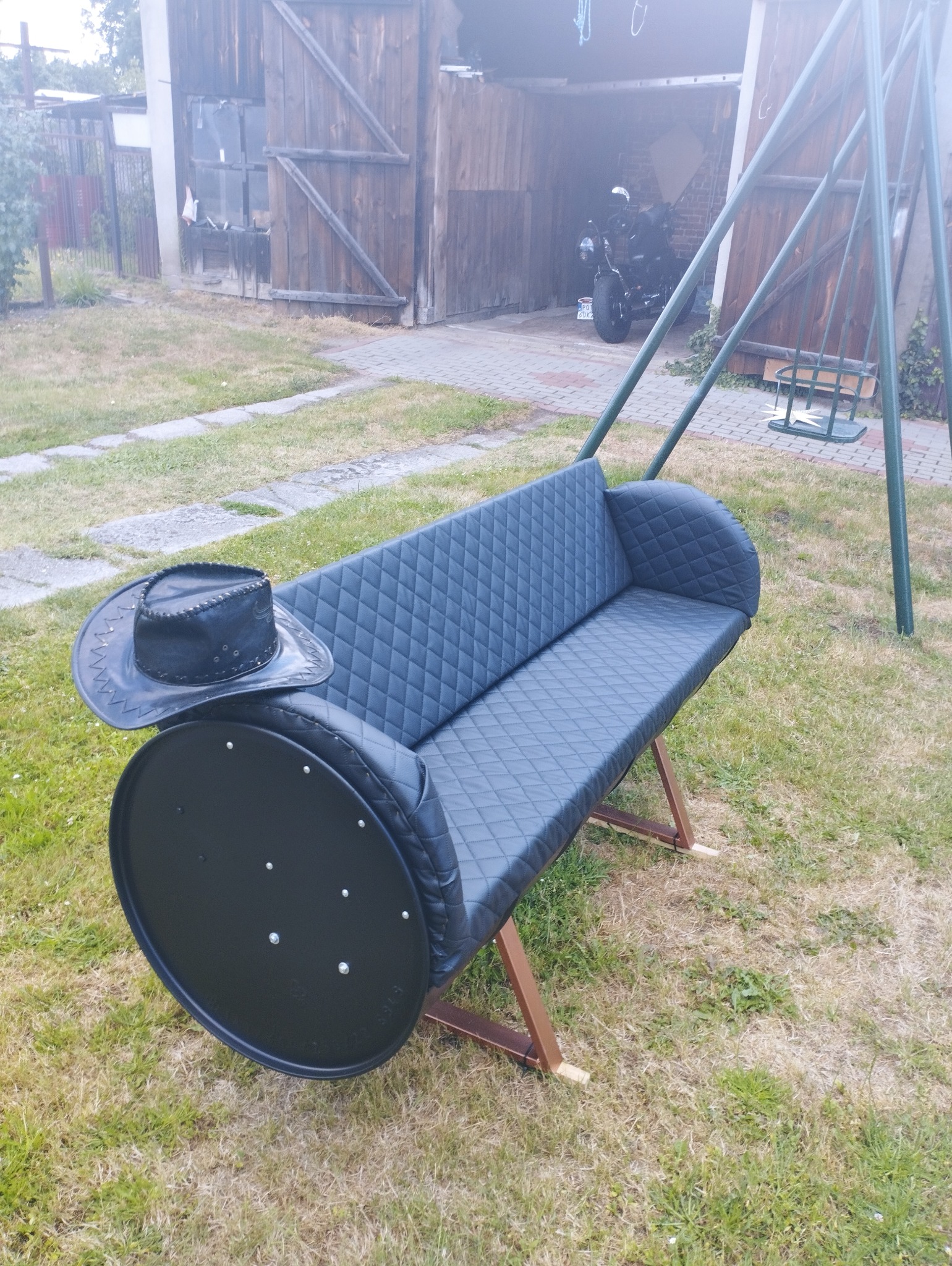 Oryginalna huśtawka-ławka wykonana z beczki i obita czarną skórą z pikowaniem w romby, z czarnym kowbojskim kapeluszem na jednym z końców, stojąca na trawniku przed garażem z motocyklem w środku.